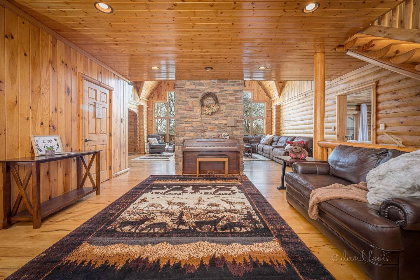 A spacious rustic living room with wood-paneled walls and ceiling, a stone fireplace at the center, large windows, leather sofas, a patterned area rug, and decorative pillows and stuffed animals.