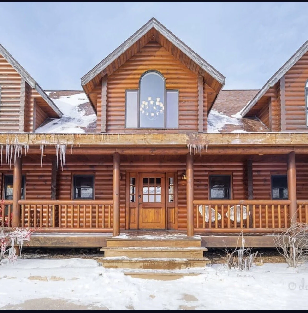 Log cabin-style house with a front porch, icicles hanging from the roof, snow on the ground, and snow on the roof. The house has large windows and a wooden door with stairs leading up to it.