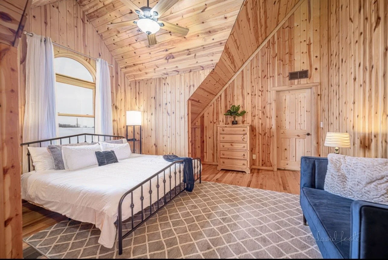 A cozy bedroom with wood-paneled walls, a metal bed frame with white bedding, a window with white curtains, a blue armchair, a beige patterned rug, and a wooden dresser with a plant on top.