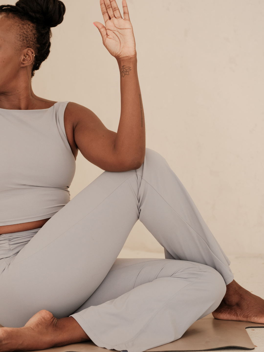 A person practicing yoga or meditation, sitting cross-legged on a mat with one arm raised and palm facing outward, wearing a light beige tank top and matching pants, with visible tattoos on the arm, against a neutral background.