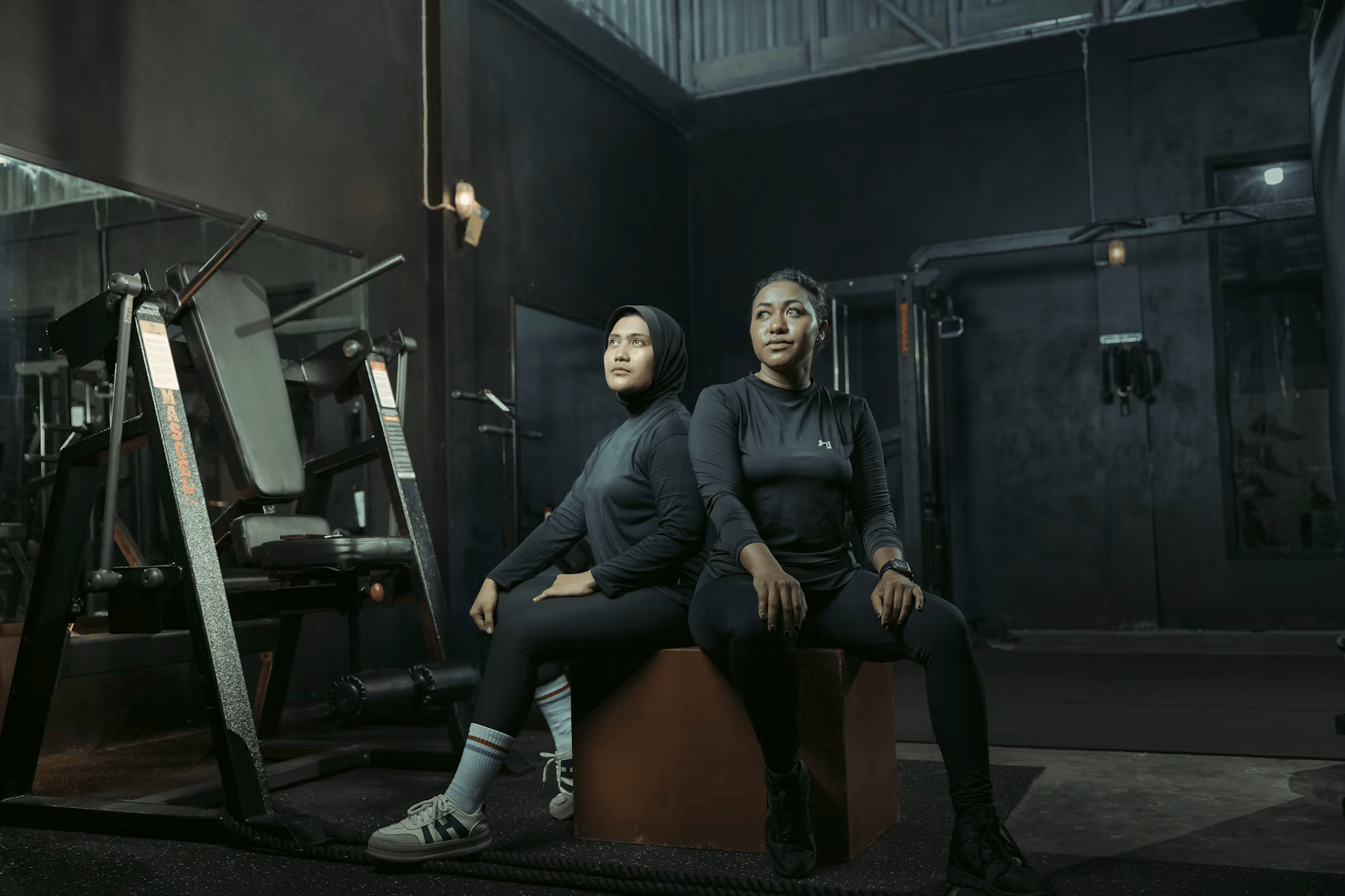 Two women in black athletic clothing sitting back-to-back on a brown box in a dark gym, with exercise equipment in the background.
