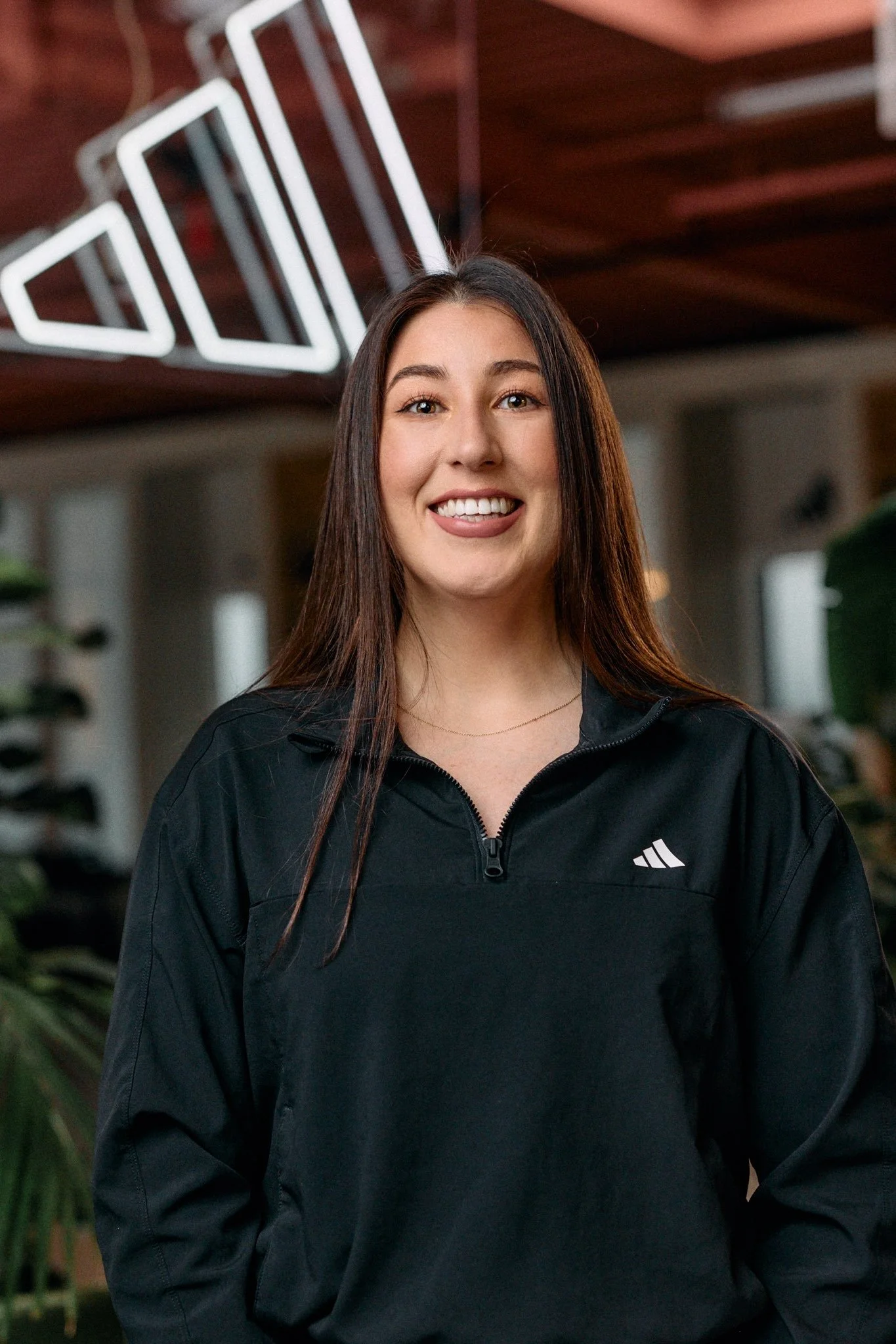 A smiling woman with long brown hair wearing a black Adidas sports jacket, standing indoors with blurred greenery and a ceiling with neon lights in the background.