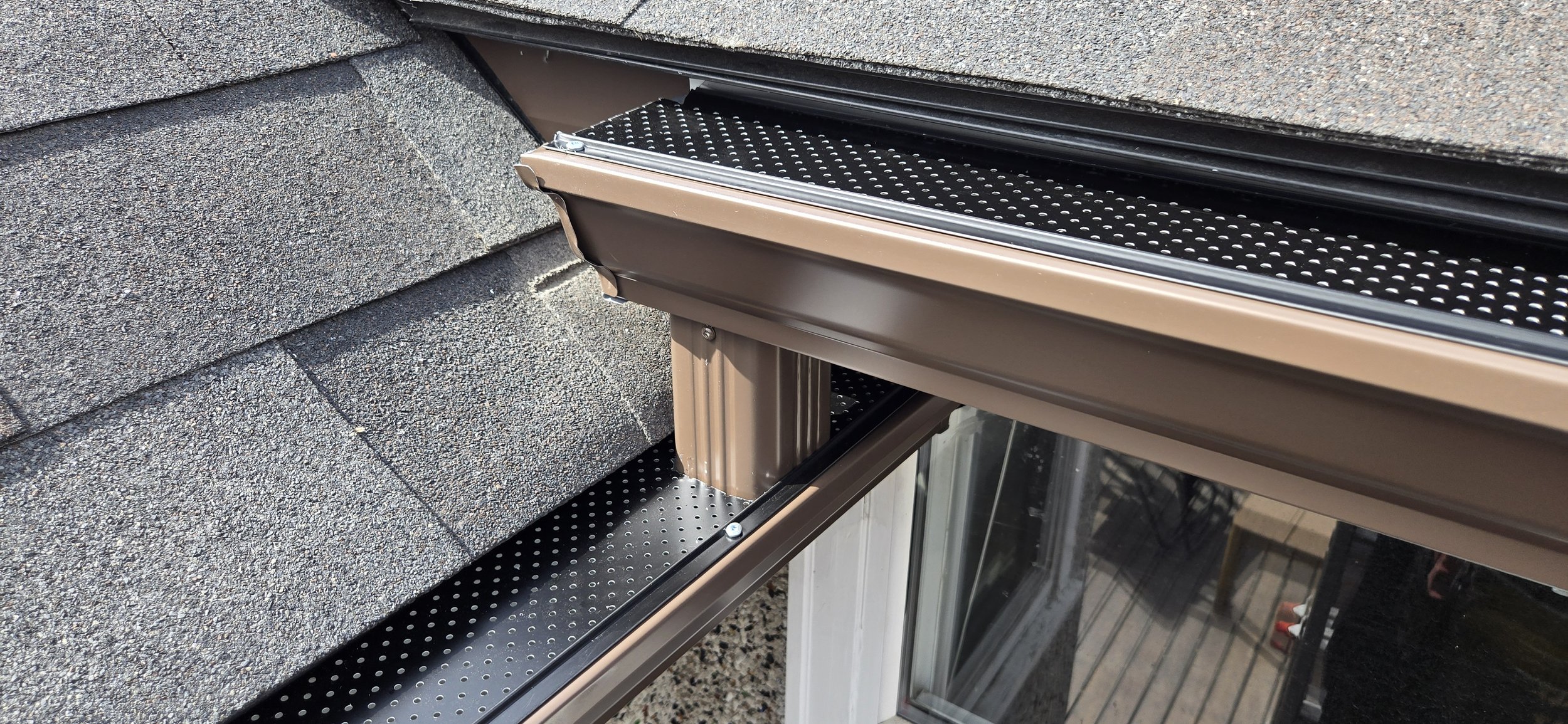 Close-up view of a house roof with textured asphalt shingles and a metal gutter system with black perforated lining.