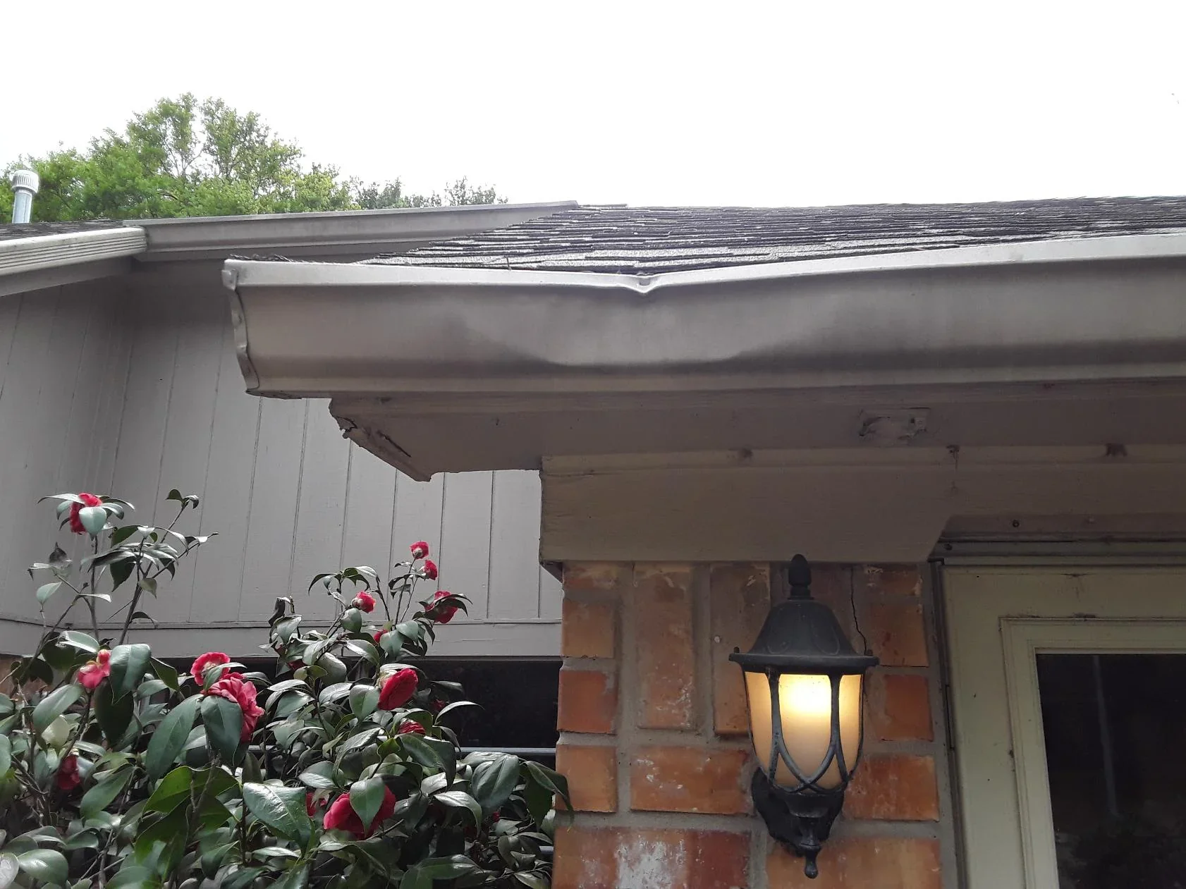 Close-up view of the corner of a house rooftop showing damaged gutter, brick wall, and outdoor lamp with warm light, with some greenery and pink flowers in the foreground.