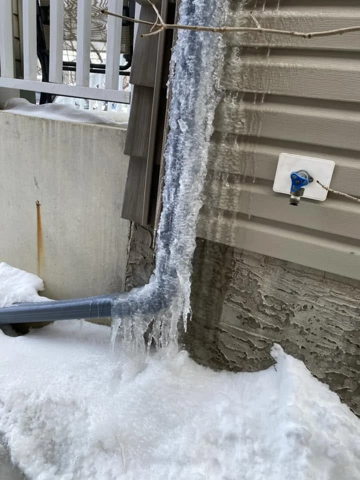 Ice formations hanging from the side of a house near a water spigot, with snow and ice accumulation on the ground.