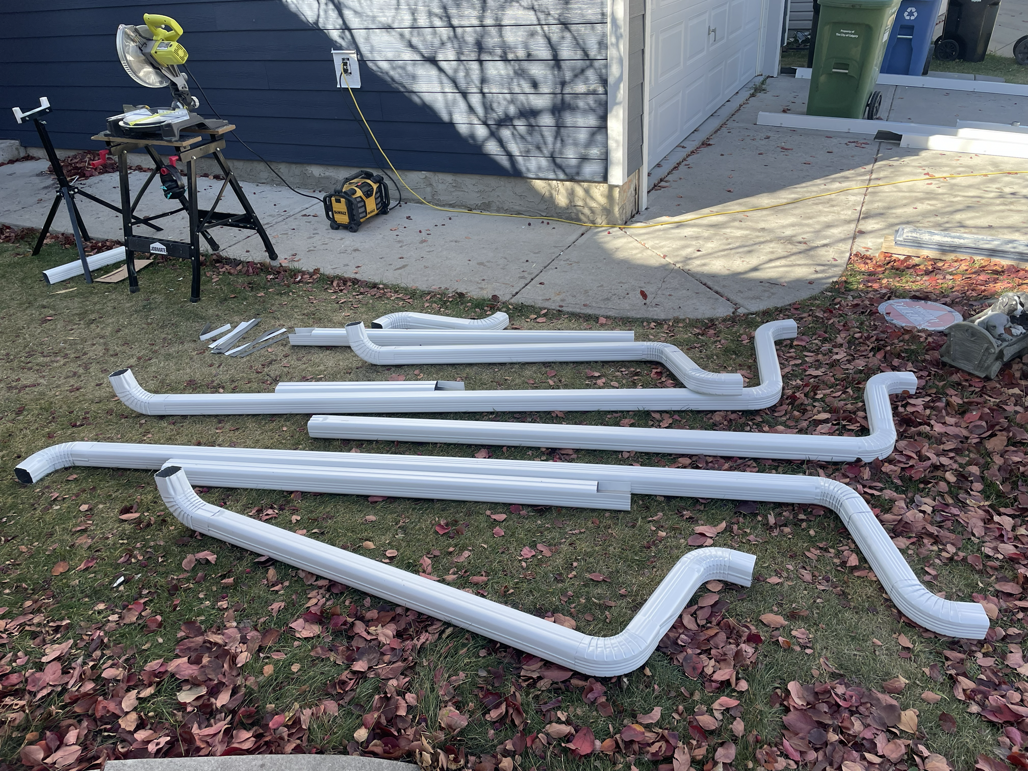Lawn with fallen leaves, construction materials, white PVC pipes, a saw on a sawhorse, and tools near a garage with blue siding.
