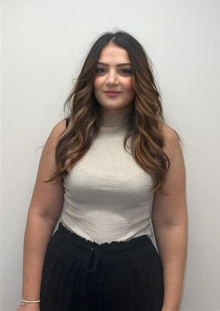A young woman with long, wavy brown hair and a neutral expression, standing against a plain white wall, wearing a sleeveless beige top and black pants.