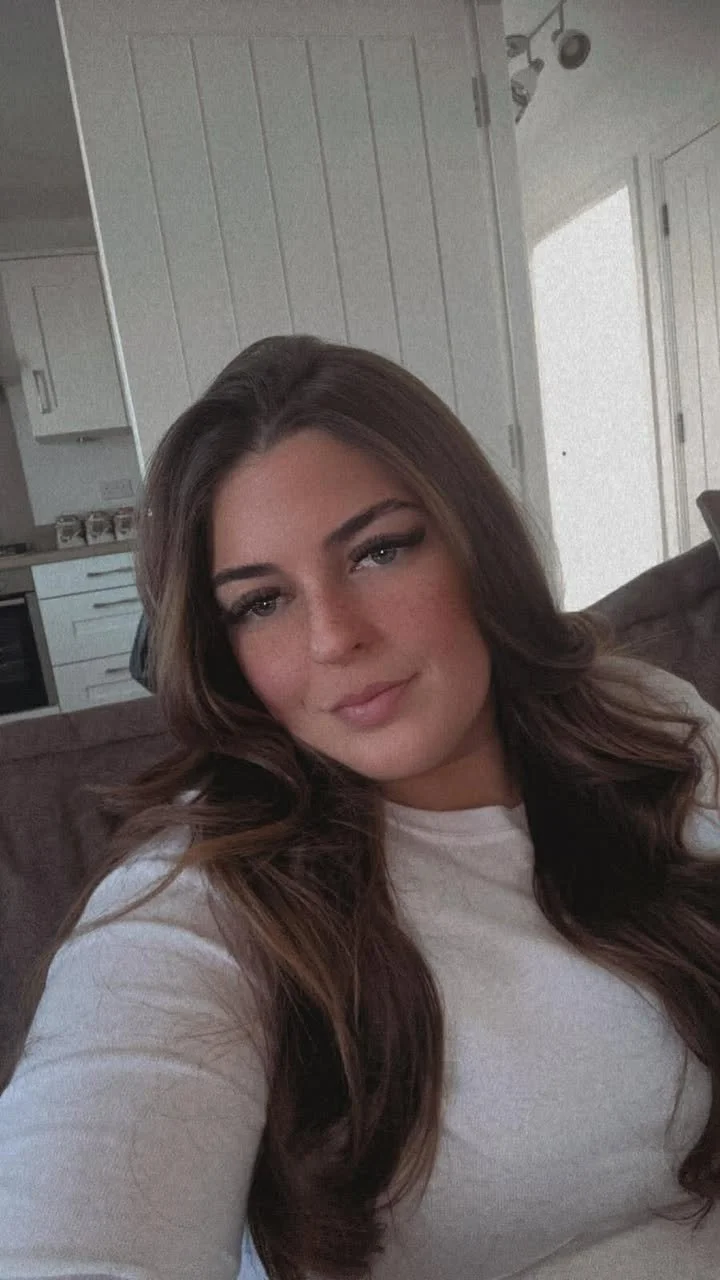 A woman with long, wavy brown hair sitting on a brown couch in a living room, with a white wall and kitchen visible in the background.