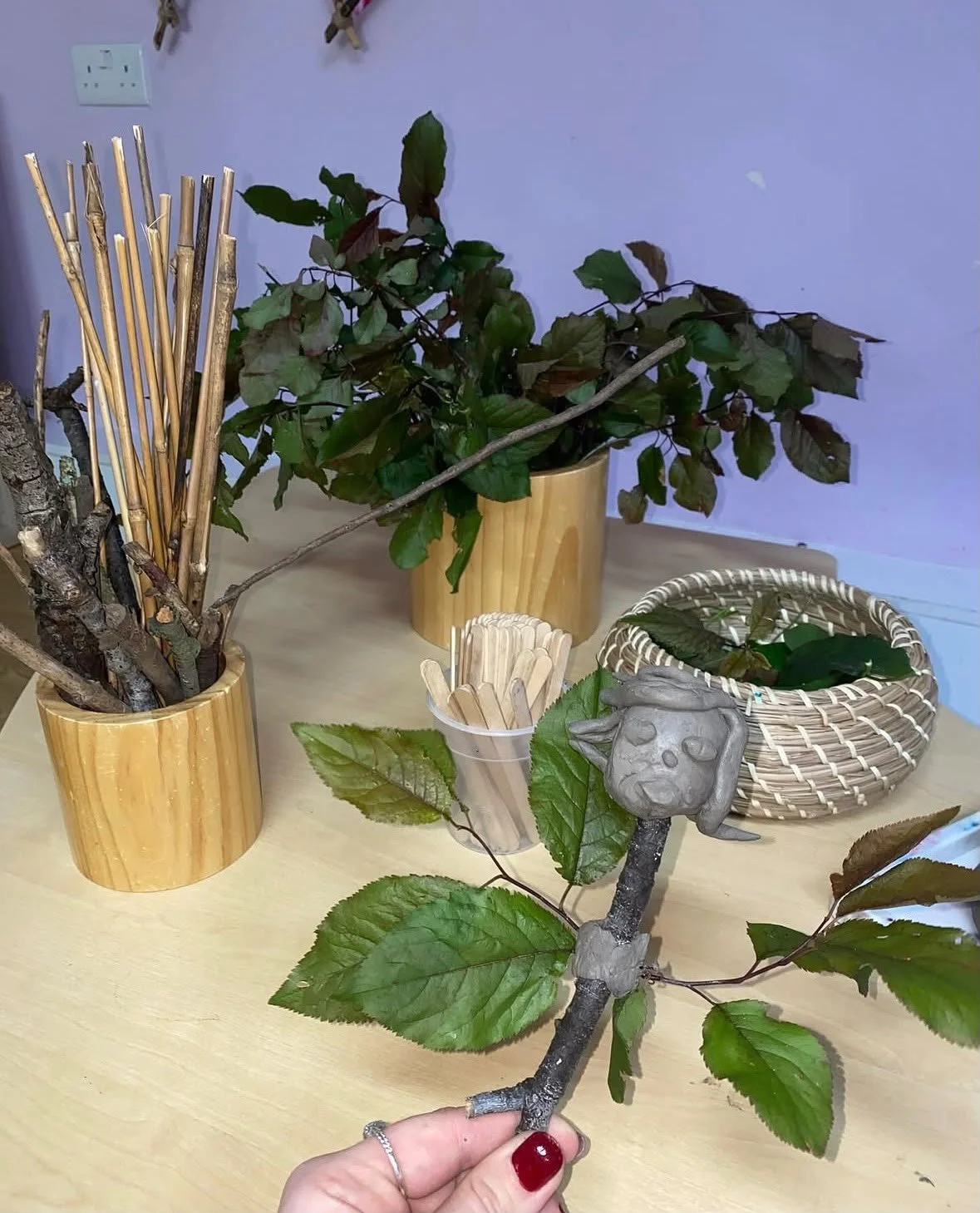 A small clay frog figurine sitting on a branch, held by a hand with red fingernail polish, surrounded by green leaves, with decorative plants and wooden sticks in containers on a light-colored table, and a purple wall in the background.
