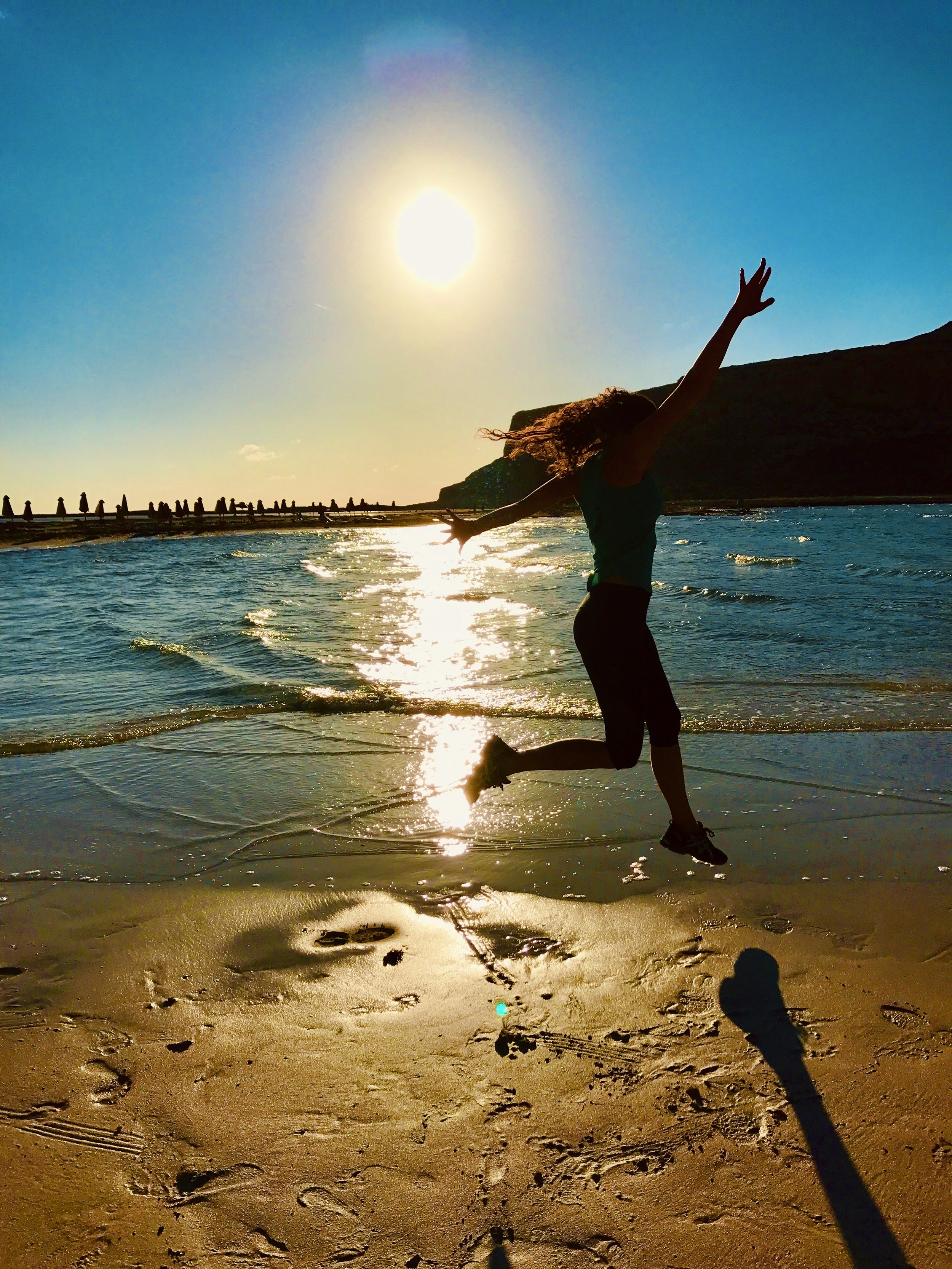 A person jumping on the beach during sunset with arms outstretched, casting a long shadow on the sand, with the ocean and cliffs in the background.