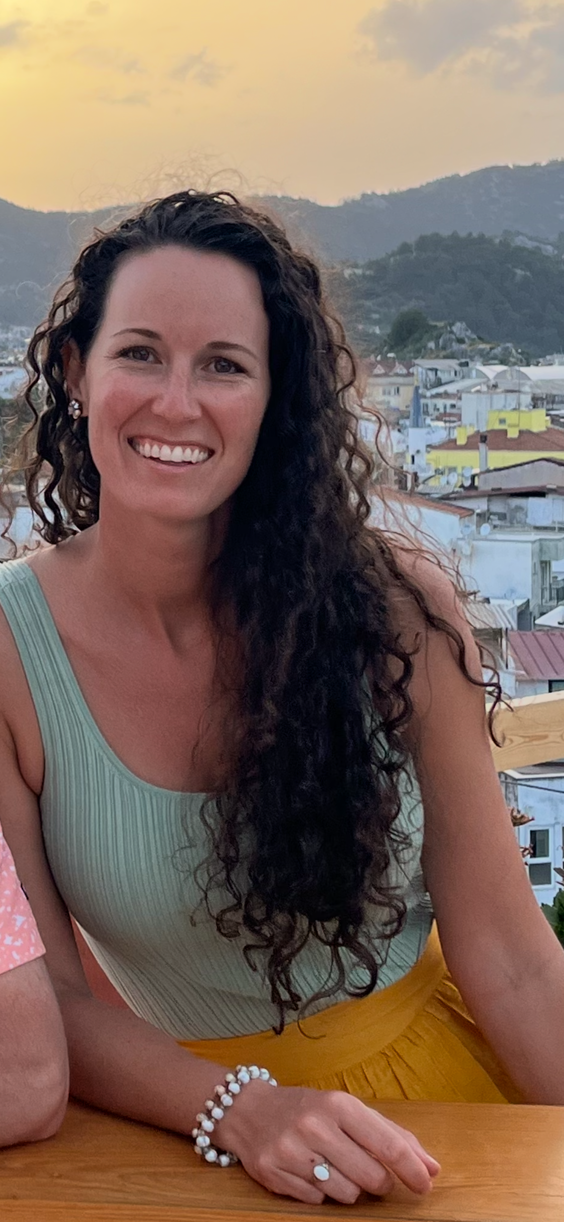 A woman with curly dark hair smiling, wearing a light green ribbed tank top, yellow skirt, and jewelry, sitting at a table with a scenic city and hills in the background during sunset.