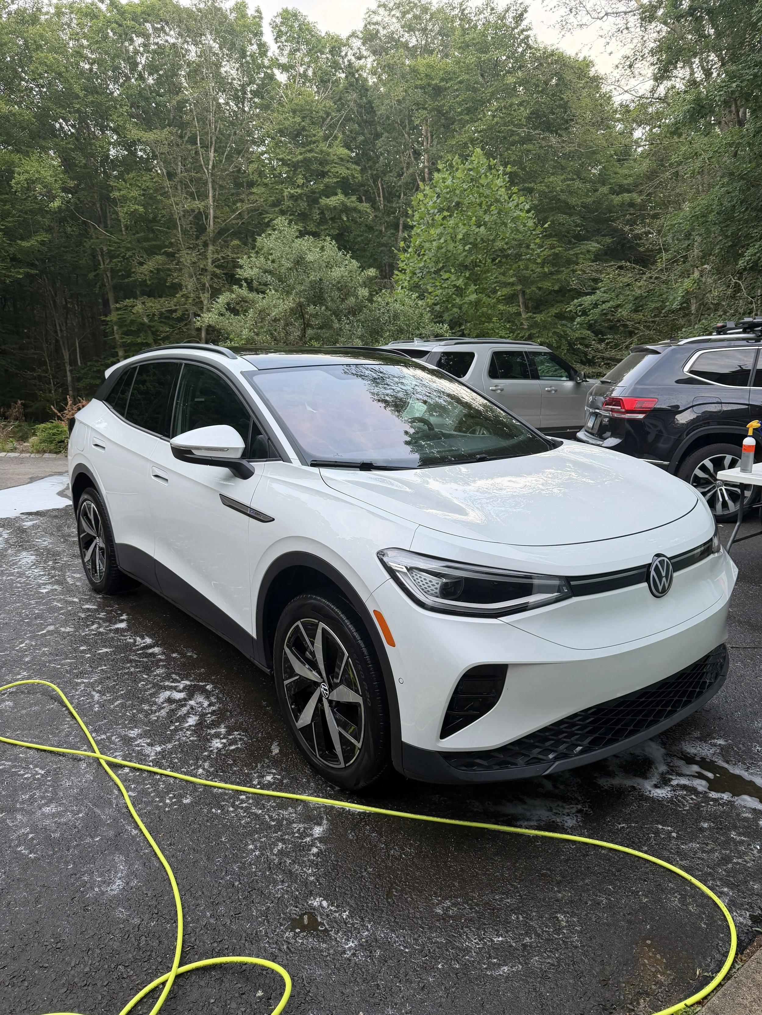 A white Volkswagen electric car parked outdoors on a wet surface, with a yellow garden hose nearby and several other parked vehicles in the background, surrounded by greenery and trees.