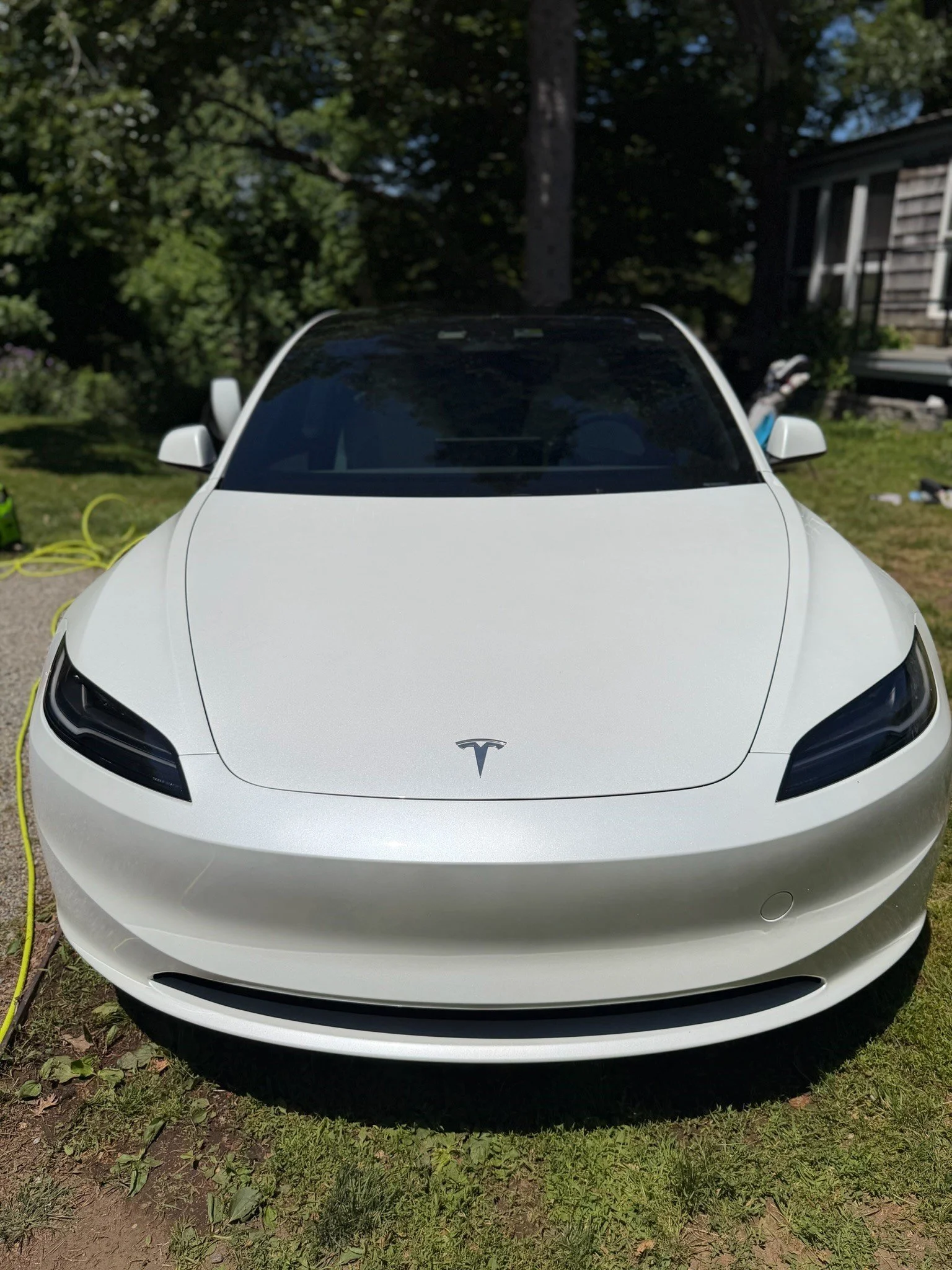 Front view of a white Tesla electric car parked on grass, with trees and a house in the background.