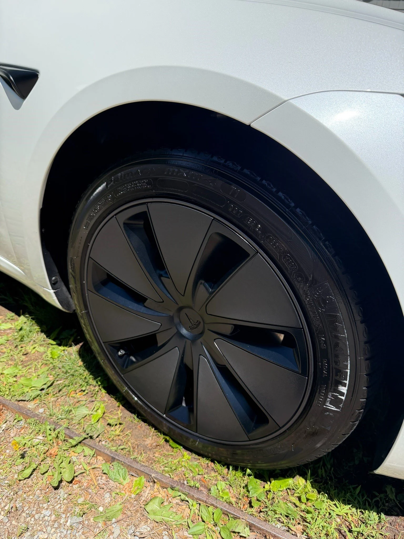 Close-up of a Tesla wheel with a black rim and tire, part of a white electric car parked on grass and gravel.