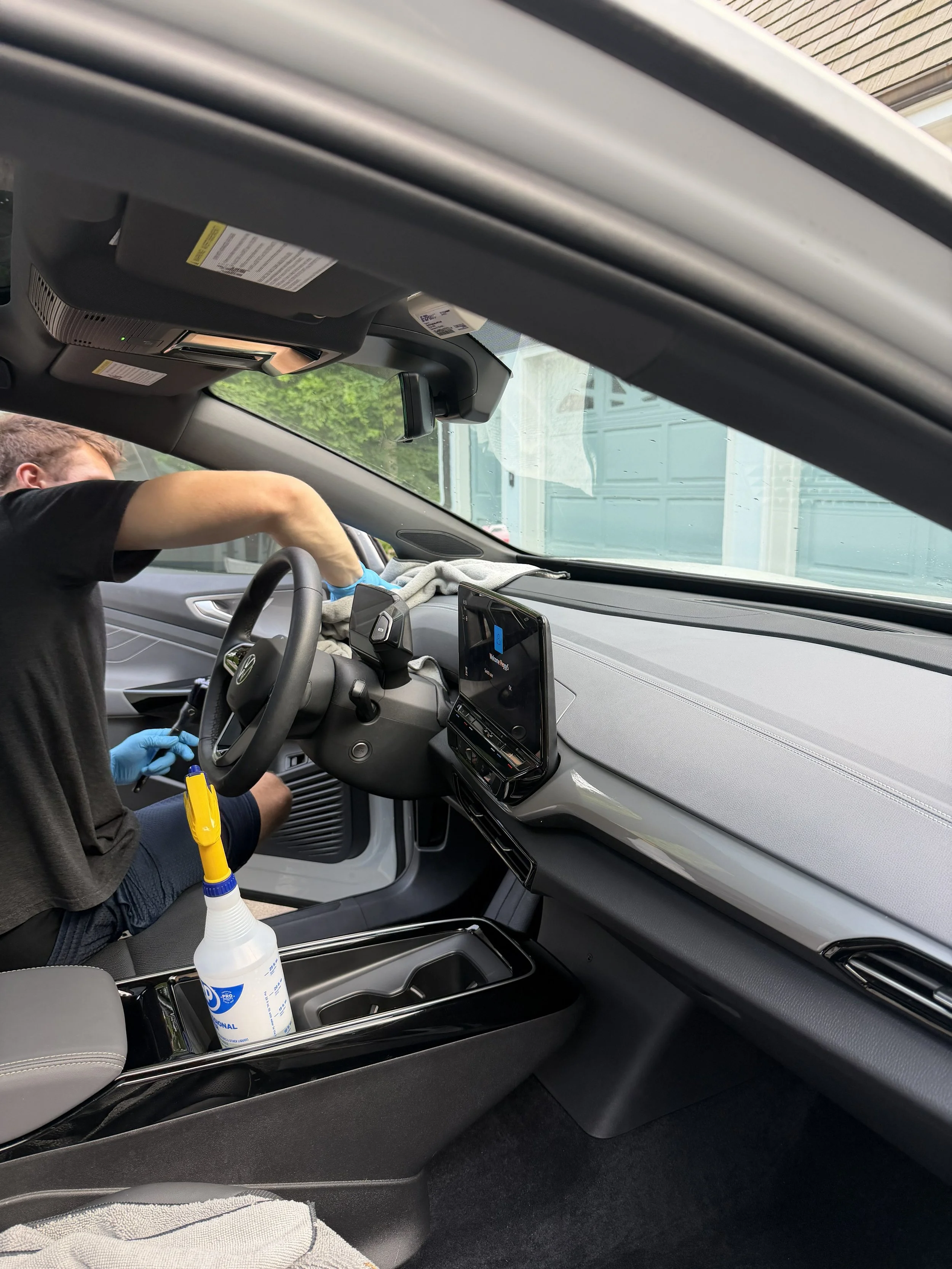 Inside a vehicle, a man is cleaning the dashboard area with a cloth. A cleaning spray bottle is in the center console.