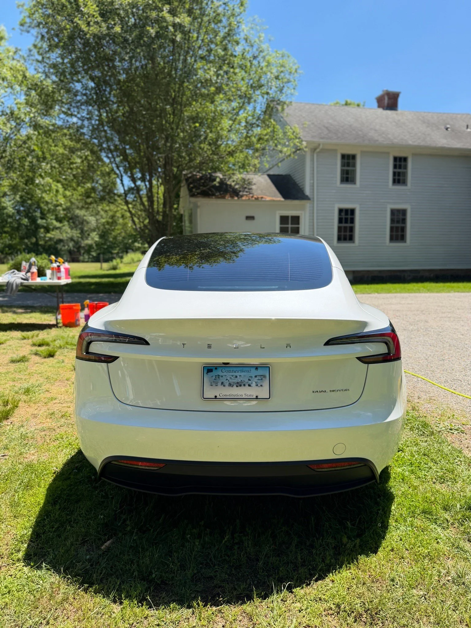 White Tesla car parked on grass in front of a white house with multiple windows and a gray roof, with trees and a blue sky in the background. A table with cleaning supplies is on the left side of the image.