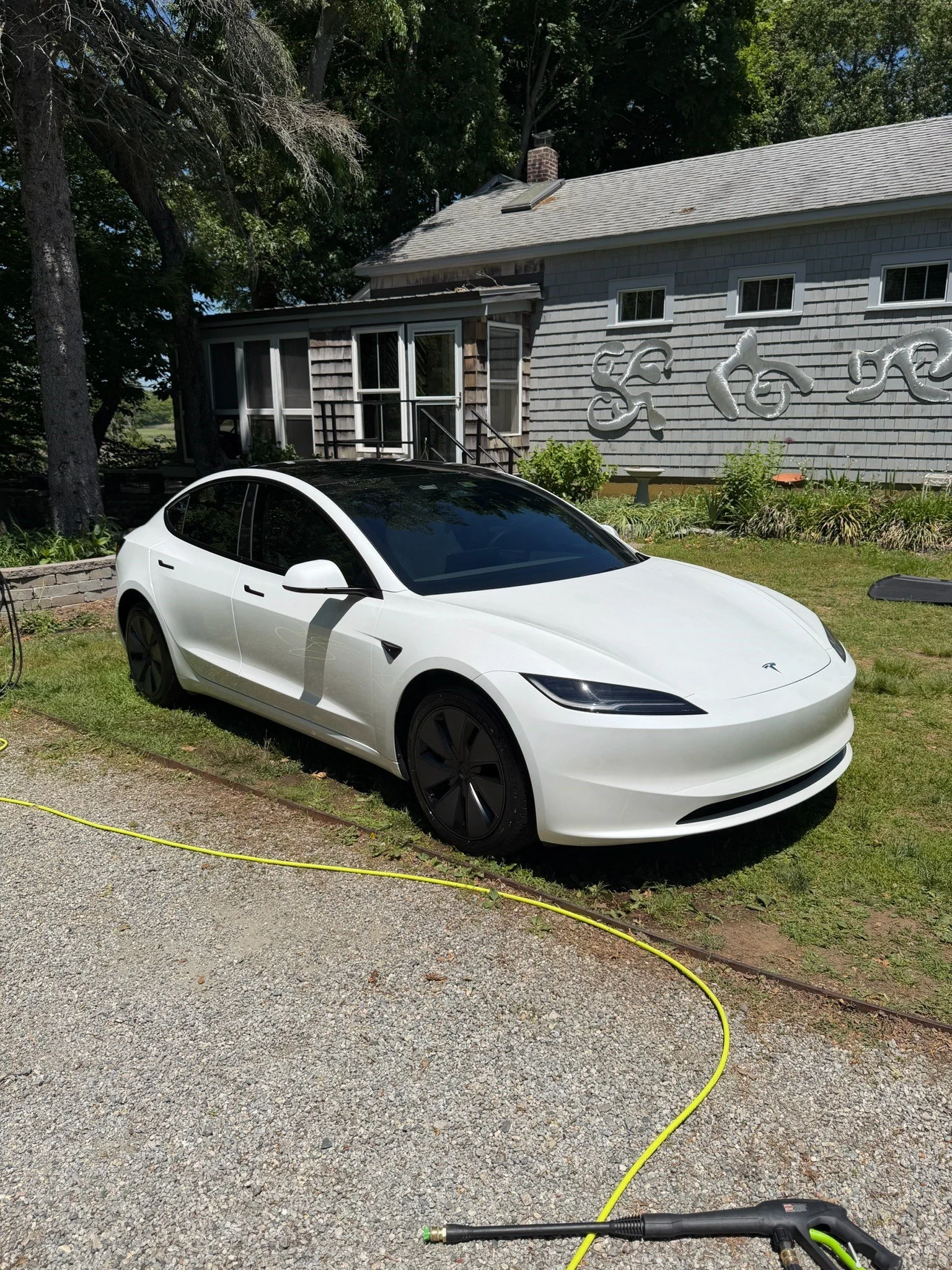 A white Tesla electric car parked on a grassy lawn in front of a house with gray shingles. A yellow extension cord is connected to the car, likely charging it. The house has multiple small windows and a porch area. There are trees and plants around t