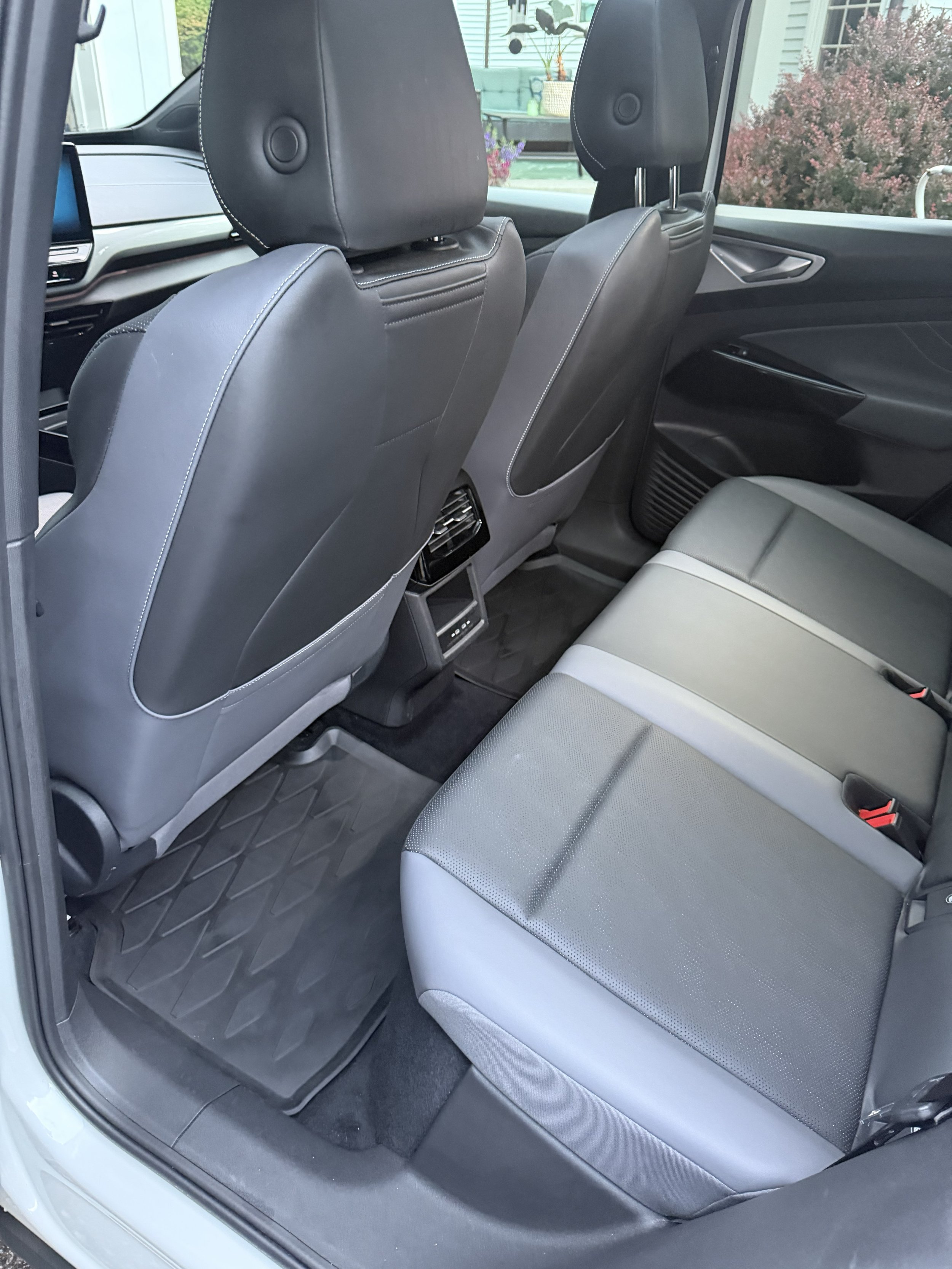 Backseat of a car showing gray upholstery, headrests on front seats, rear air vents, and a window with outdoor greenery and house in the background.