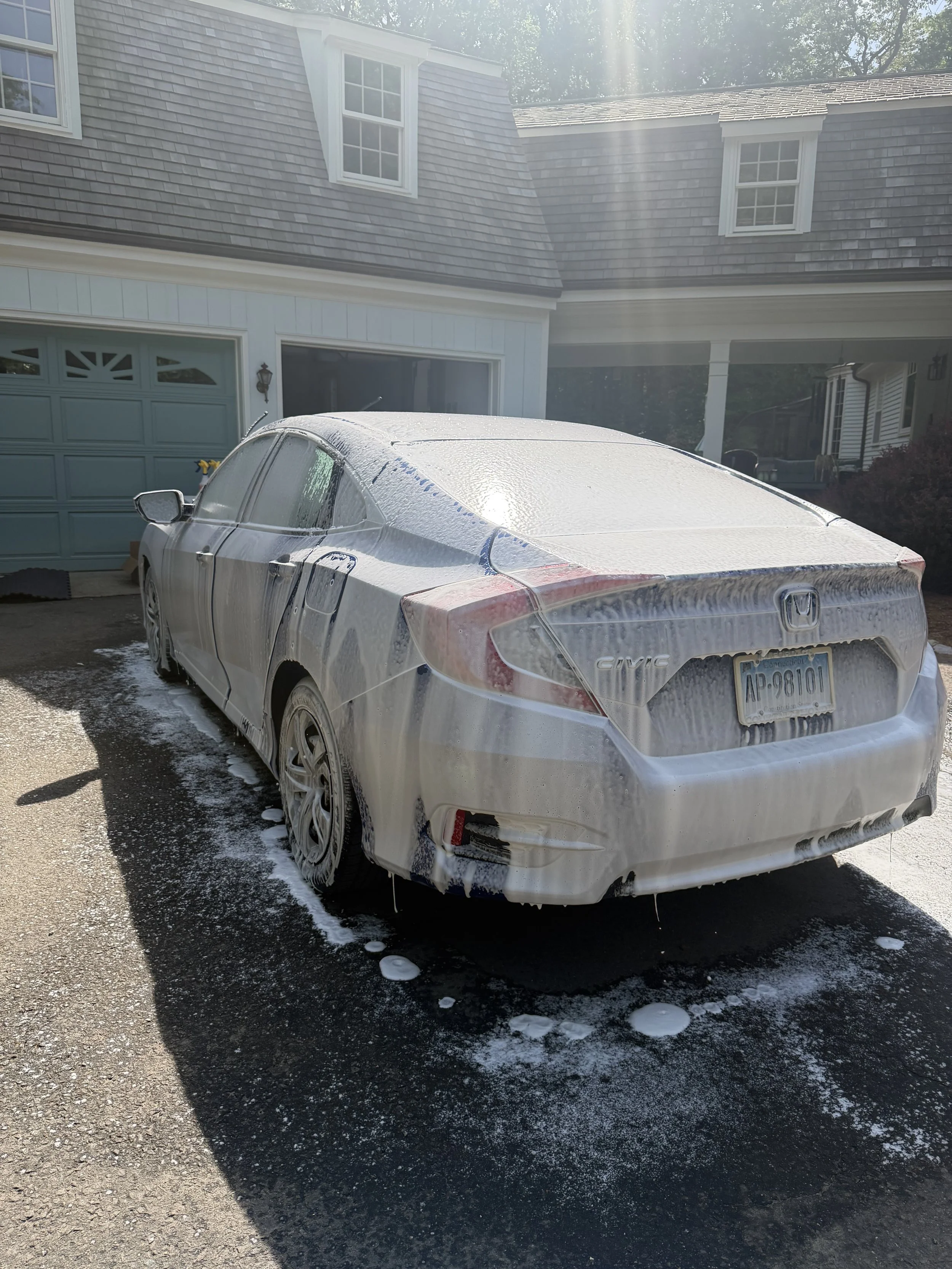 A silver Honda Civic covered in soapy foam parked in a driveway in front of a house with a blue garage door and multiple windows. Sunlight shines down, creating streaks of light in the image.