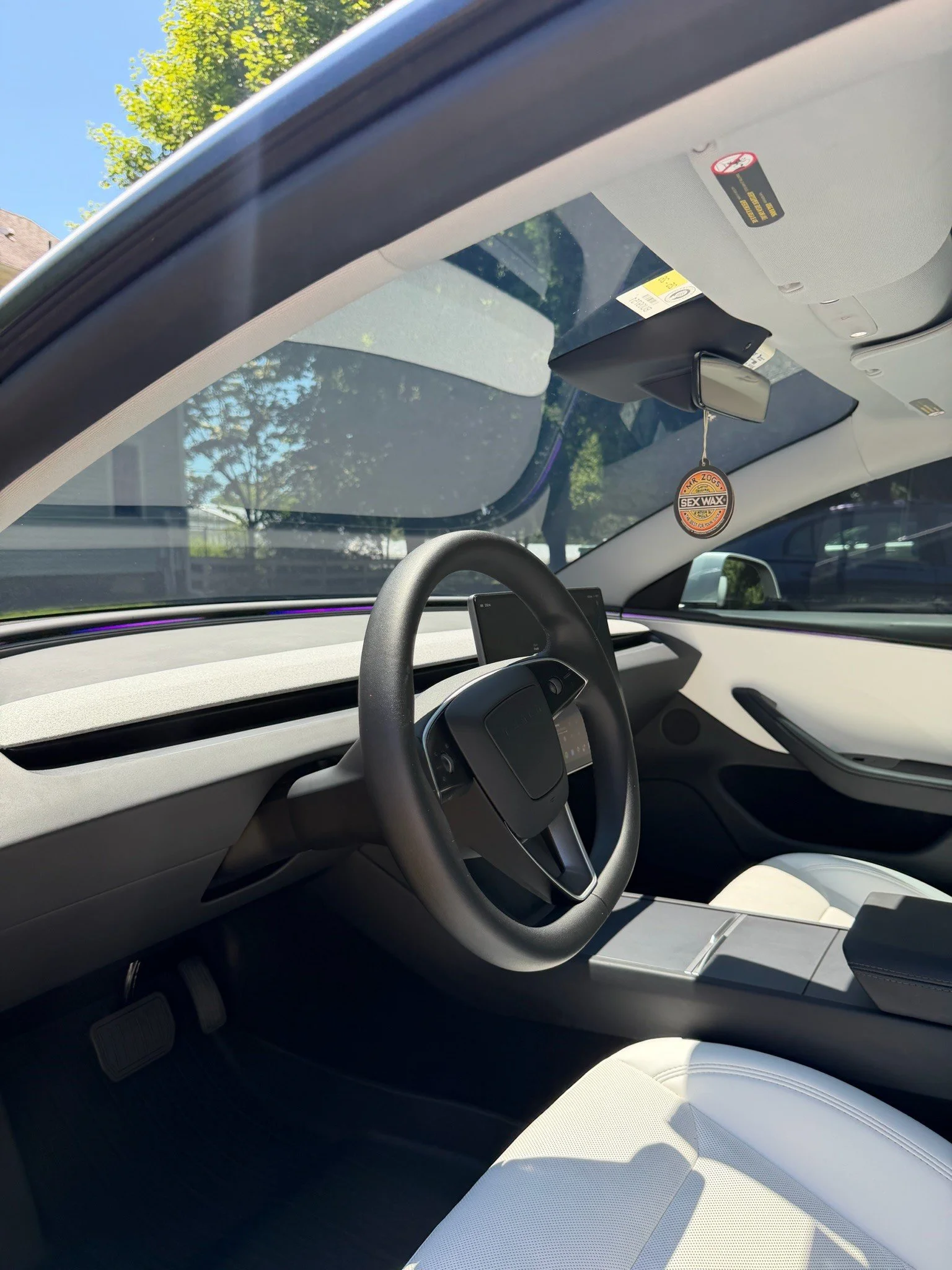 Inside a modern electric vehicle, showing the steering wheel, dashboard, and a white leather seat. The windshield reveals a tree and blue sky outside.