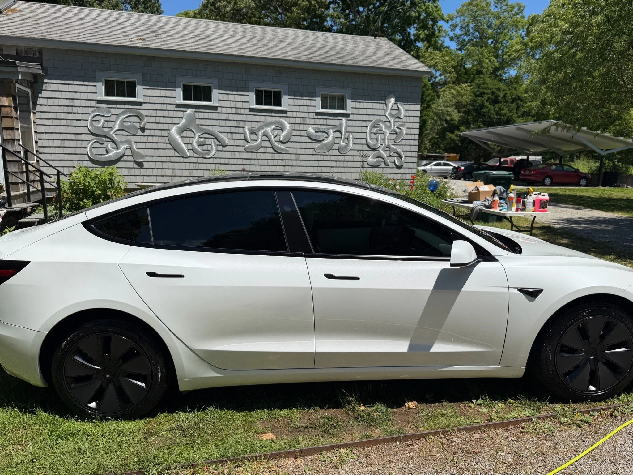 A white Tesla Model Y parked in front of a gray house with gray shingle siding. There is a table with various bottles and containers on the right side and a carport with cars behind it. Trees and a clear blue sky are in the background.