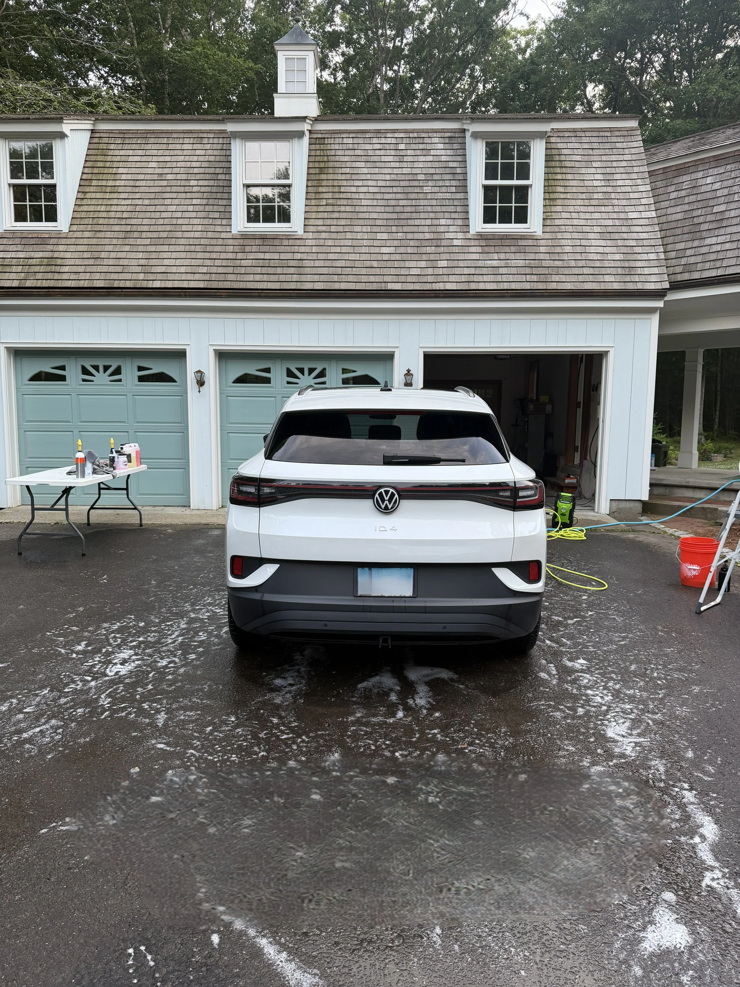 A white Volkswagen ID.4 electric SUV parked in a driveway in front of a house with blue garage doors, a partly open garage, and a wet asphalt surface with patches of snow or ice.
