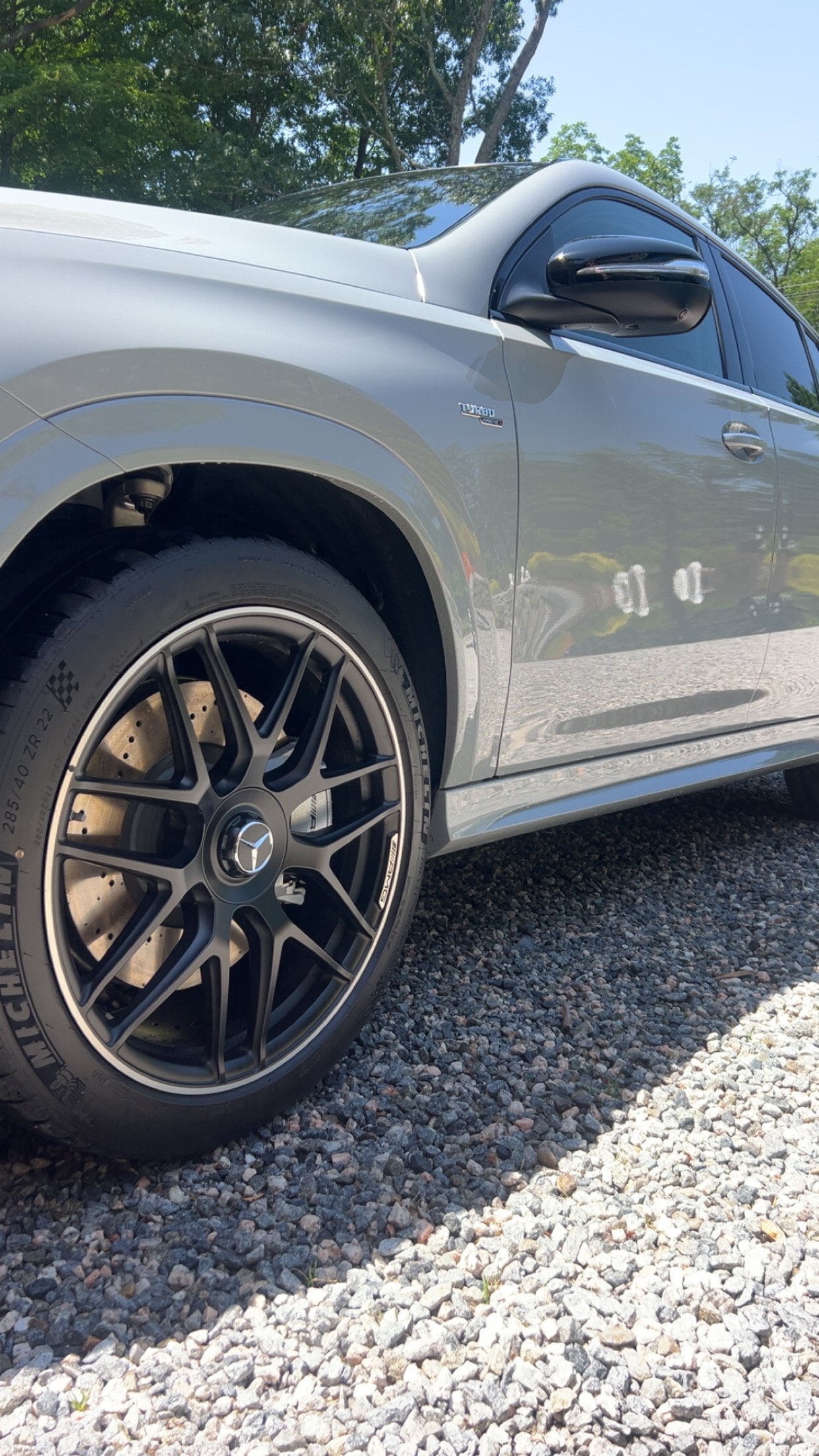 Close-up of a silver Mercedes-Benz car with black wheels, parked on a gravel surface, with trees and clear sky reflected on the car's surface.