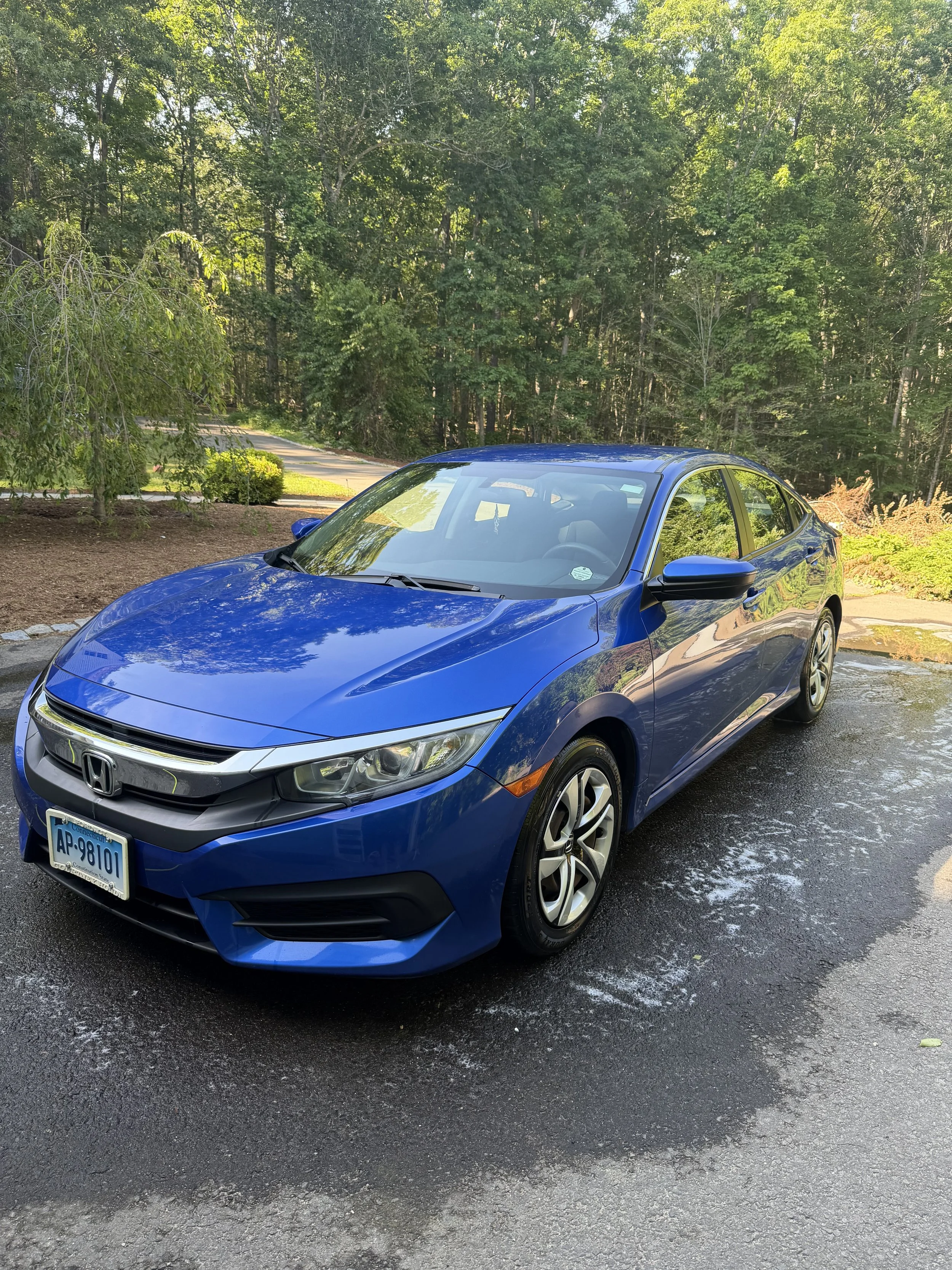 Blue Honda sedan parked on a wet asphalt surface with green trees and a small grassy area in the background.