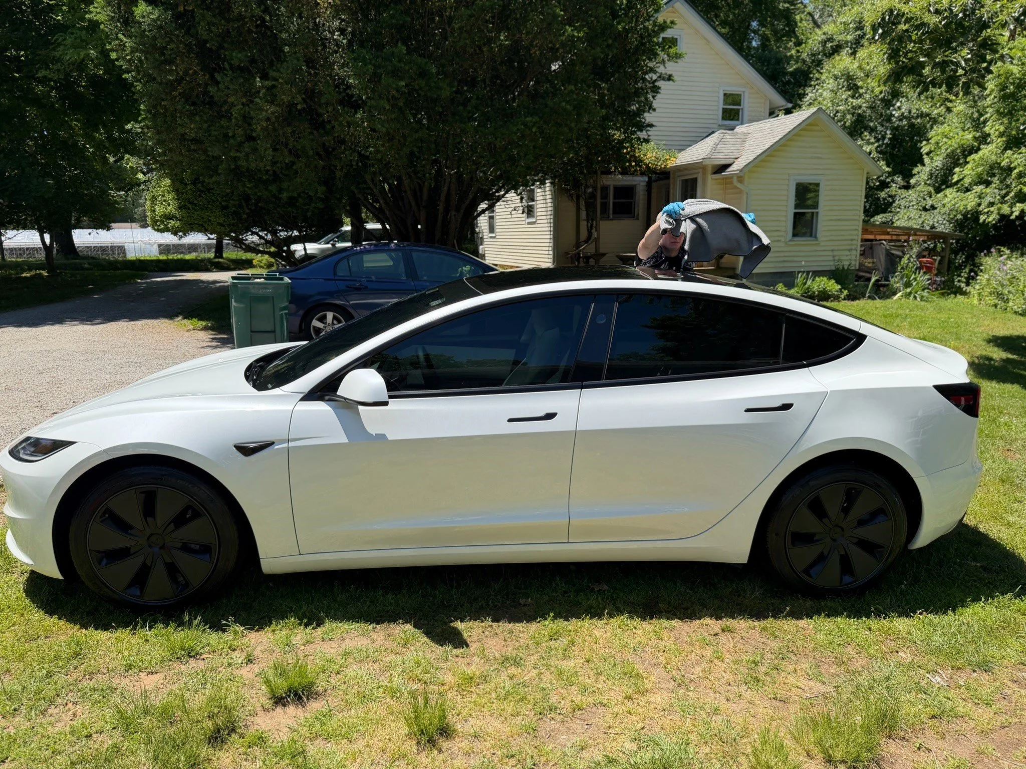 A white Tesla parked on a grassy area in front of a yellow house with a large tree nearby. A person is placing clothes into the car's trunk, wearing gloves.