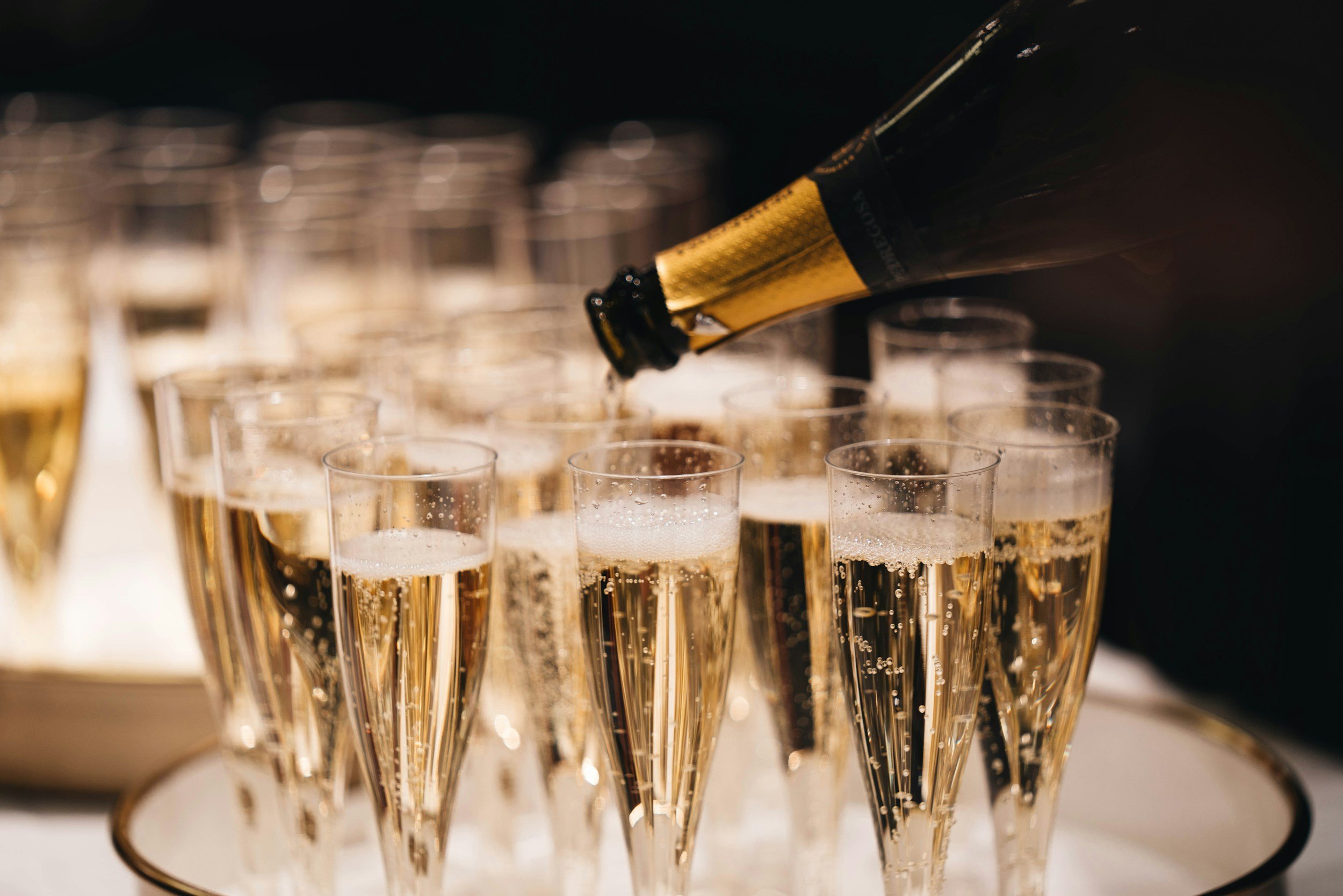 A hand pouring champagne from a bottle into tall flute glasses arranged in a row.