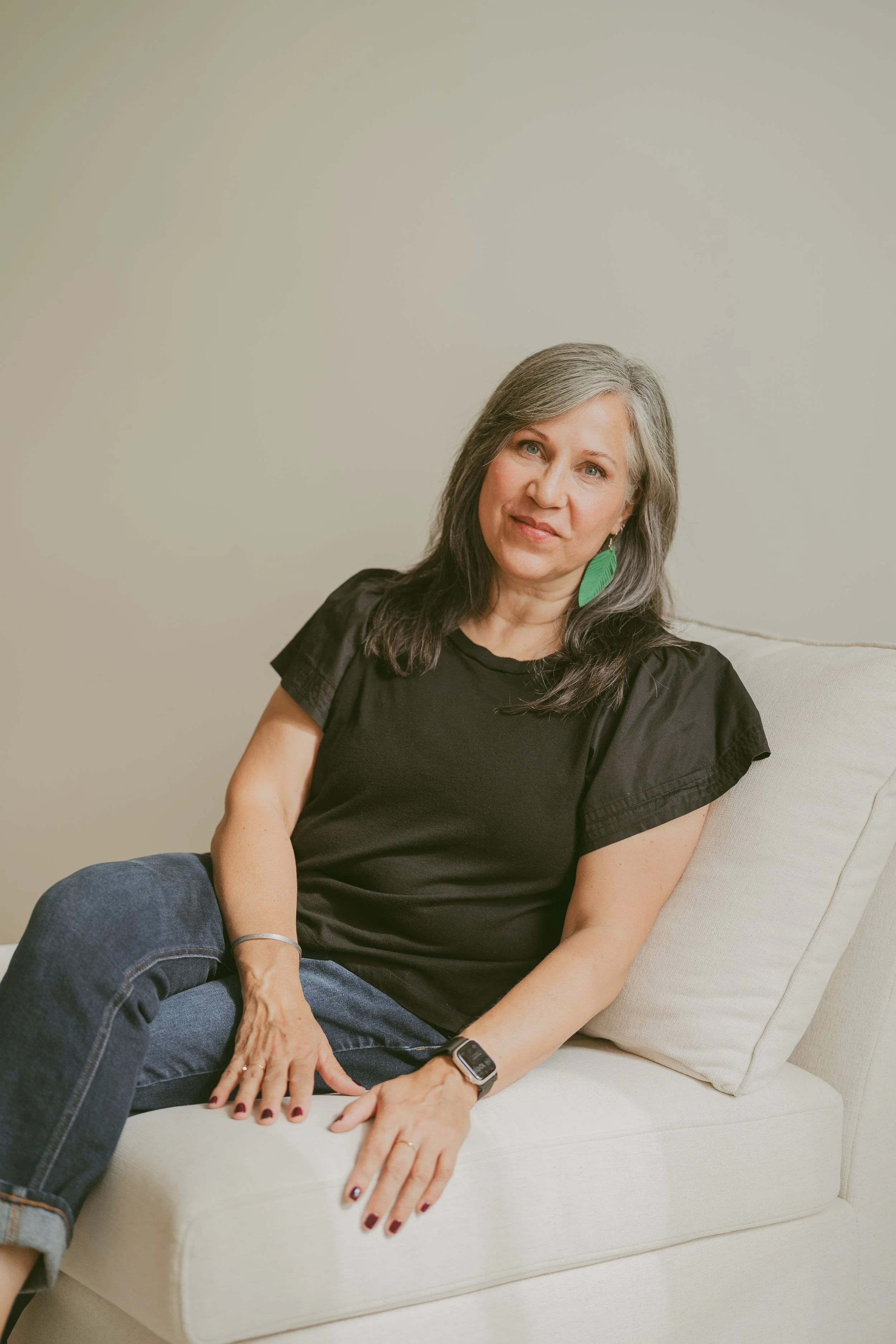 A woman with gray hair wearing a black t-shirt and jeans, sitting on a light-colored couch, looking at the camera with a slight smile, against a plain beige wall, with a green feather earring visible in her left ear.