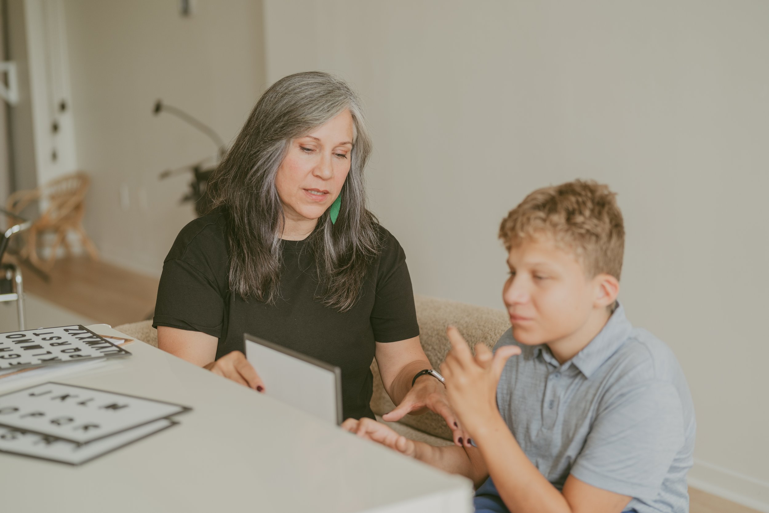 A woman with gray and black hair, green earrings, and a black shirt sits at a table with a young boy with curly hair and a gray shirt. They are engaged in a learning activity with laminated sheets of alphabet and number flashcards. The woman is looking down at the flashcards while the boy is smiling and gesturing with his hands.