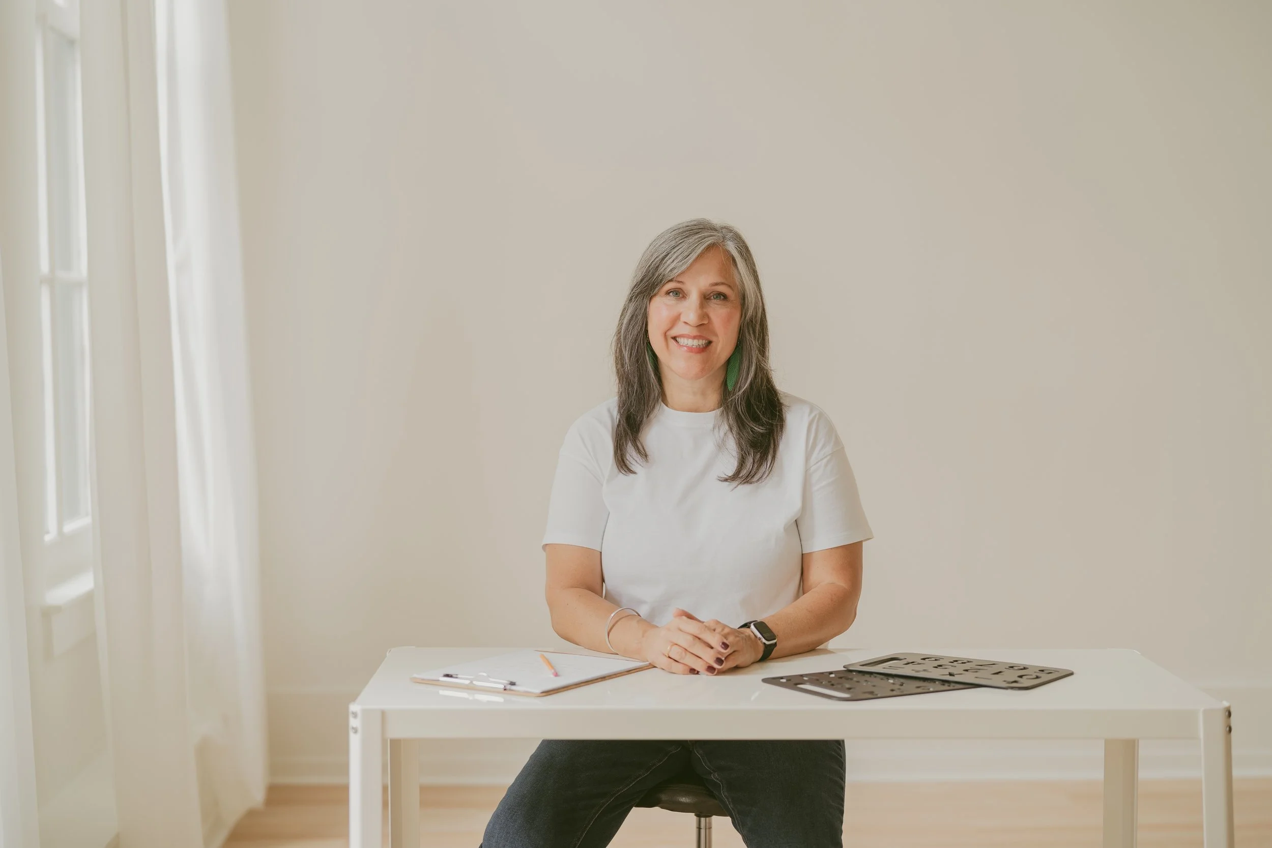 A woman with gray hair wearing a white t-shirt sitting at a white desk in a bright room. On the desk are a clipboard, a pencil, a cutting stencil, and some papers. She is smiling and looking at the camera.
