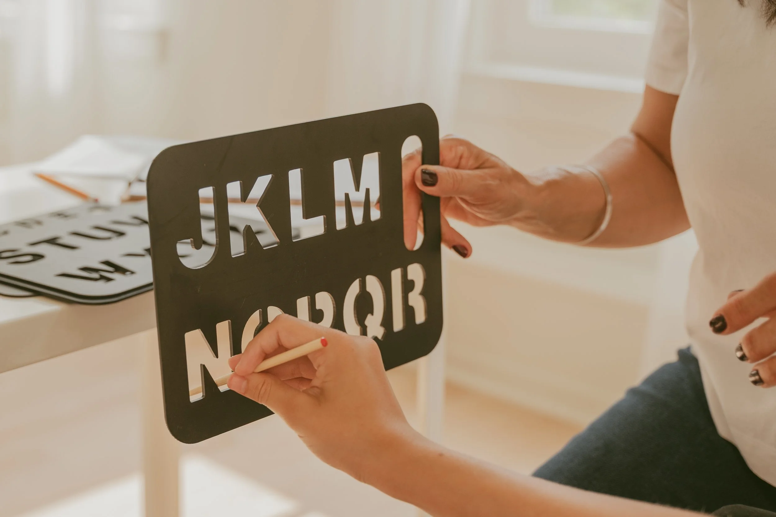 A person holding a black stencil with alphabet letters cut out. Someone else is tracing the letters onto a white surface with a small paintbrush.