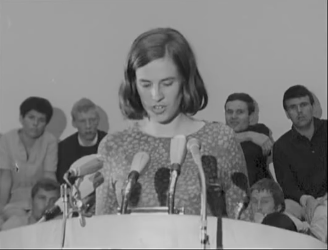 A woman with shoulder-length hair at a podium speaking into multiple microphones, with four women and two men seated behind her during a press conference or public speaking event.
