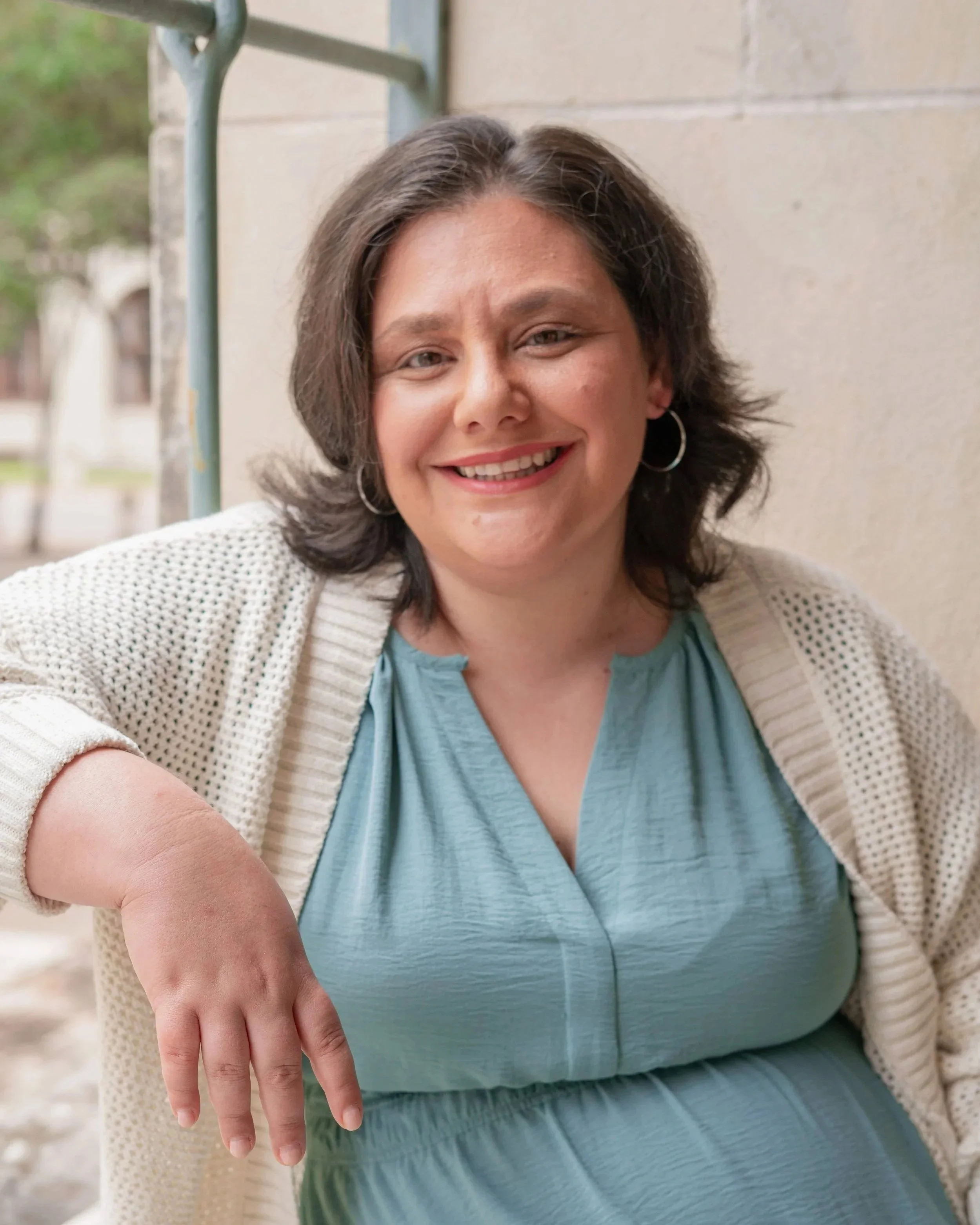 A woman with short brown hair smiling, wearing a light blue shirt, a beige cardigan, and hoop earrings, pointing at her arm where she appears to have a medical injection site.