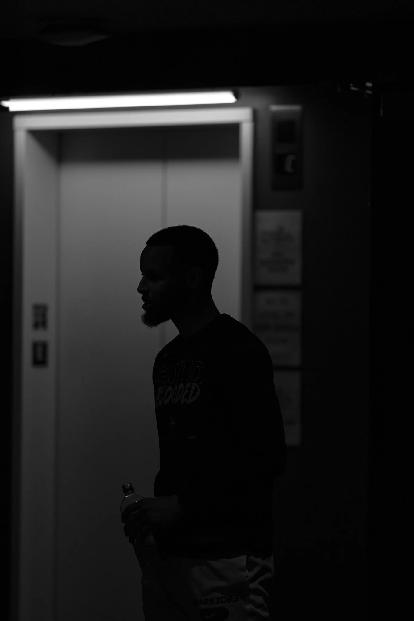 Silhouette of a man holding a water bottle in an indoor setting with a door and illuminated switch panel in the background.