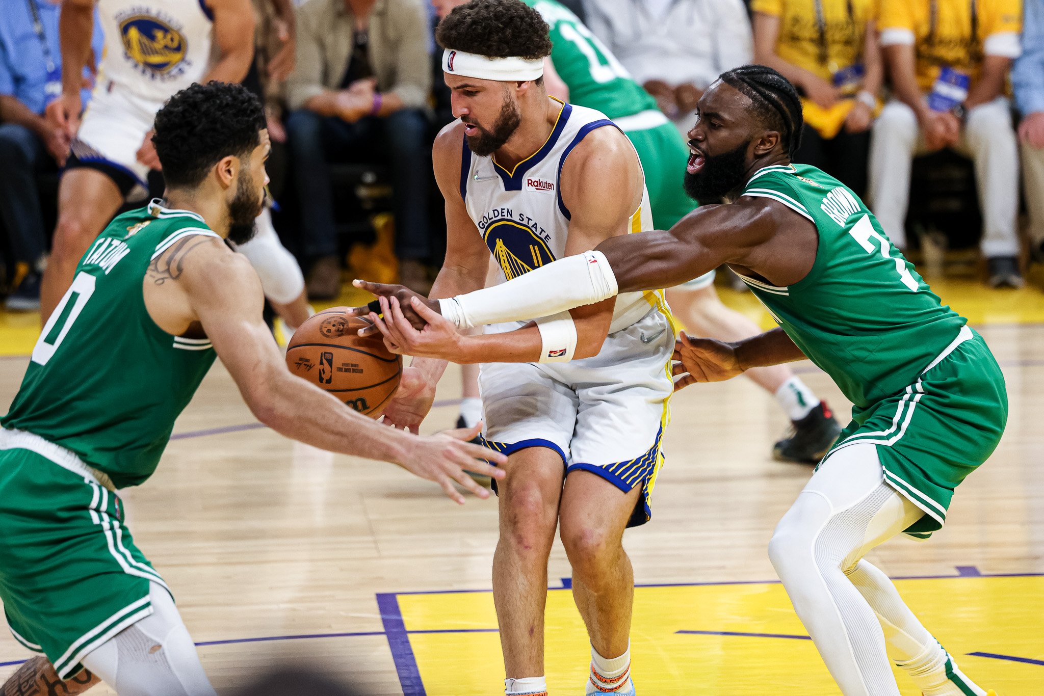 Basketball game with two players from the Golden State Warriors competing for possession of the ball against two players from the opposing team in green jerseys. The scene is on an indoor basketball court with spectators in the background.