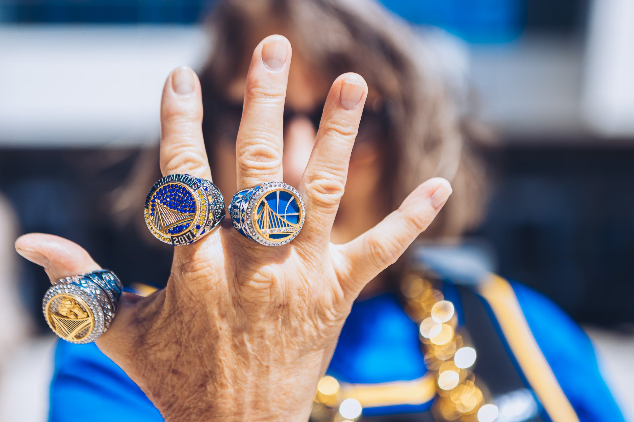 Close-up of a woman's hand with multiple basketball championship rings, with her face and hair slightly blurred in the background.