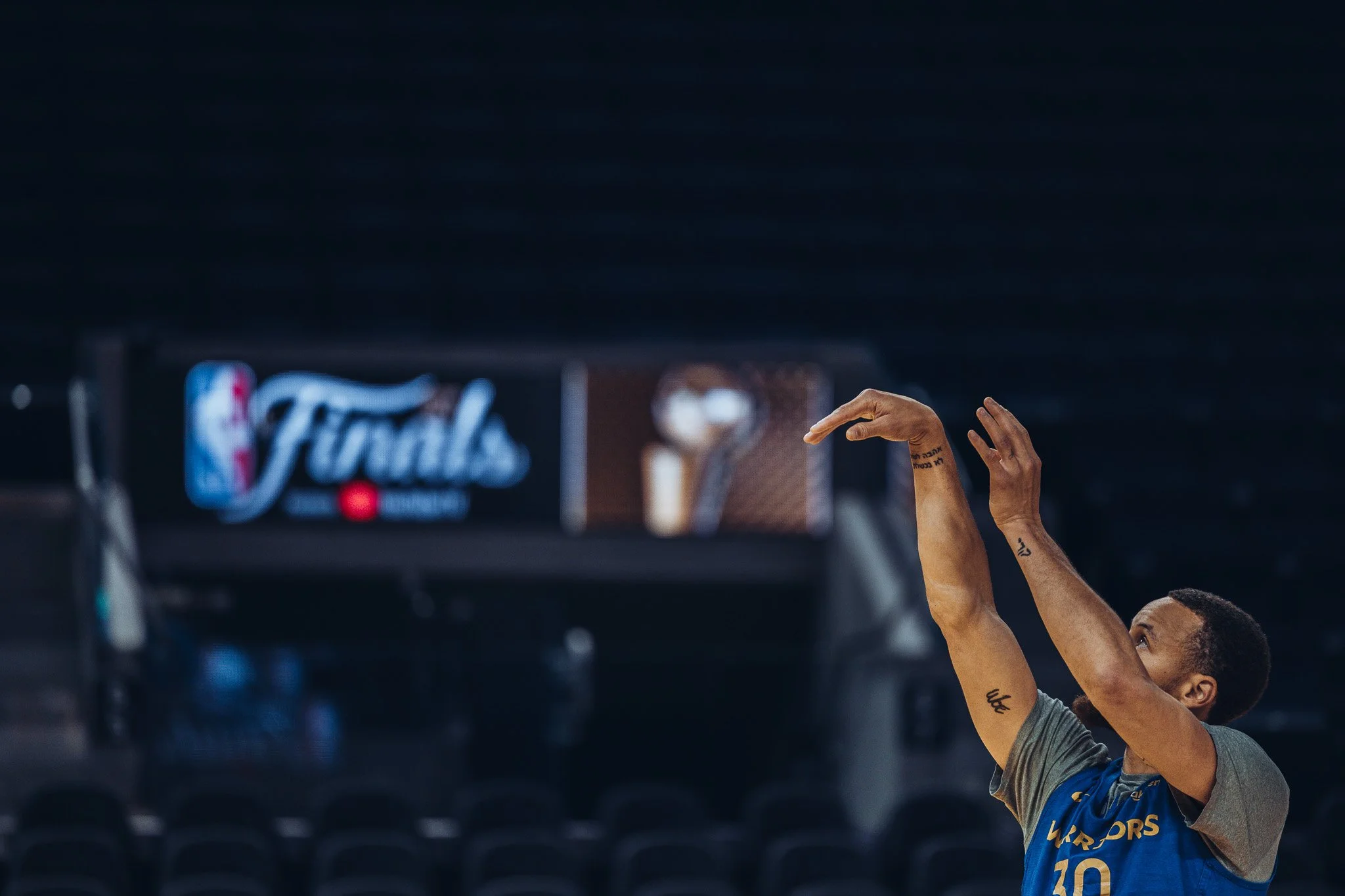 A basketball player from the Golden State Warriors is shooting a free throw during a game. The player's hands and part of their face are visible, with a scoreboard or advertisement in the background.