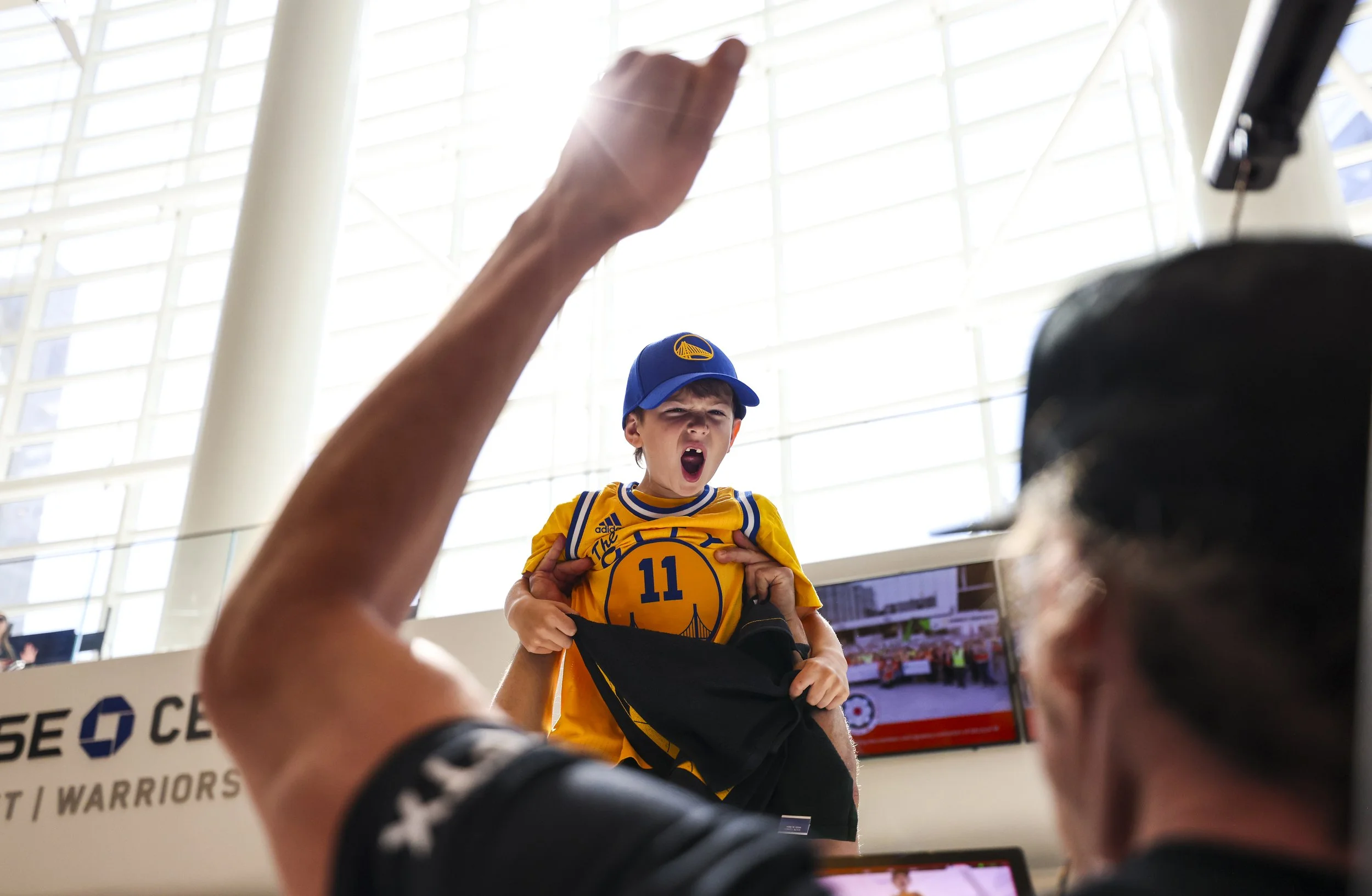 A young boy wearing a Golden State Warriors jersey and cap is being lifted in the air by someone, with his mouth open as if shouting or cheering. The background shows a large, modern indoor space with glass walls and bright lighting.