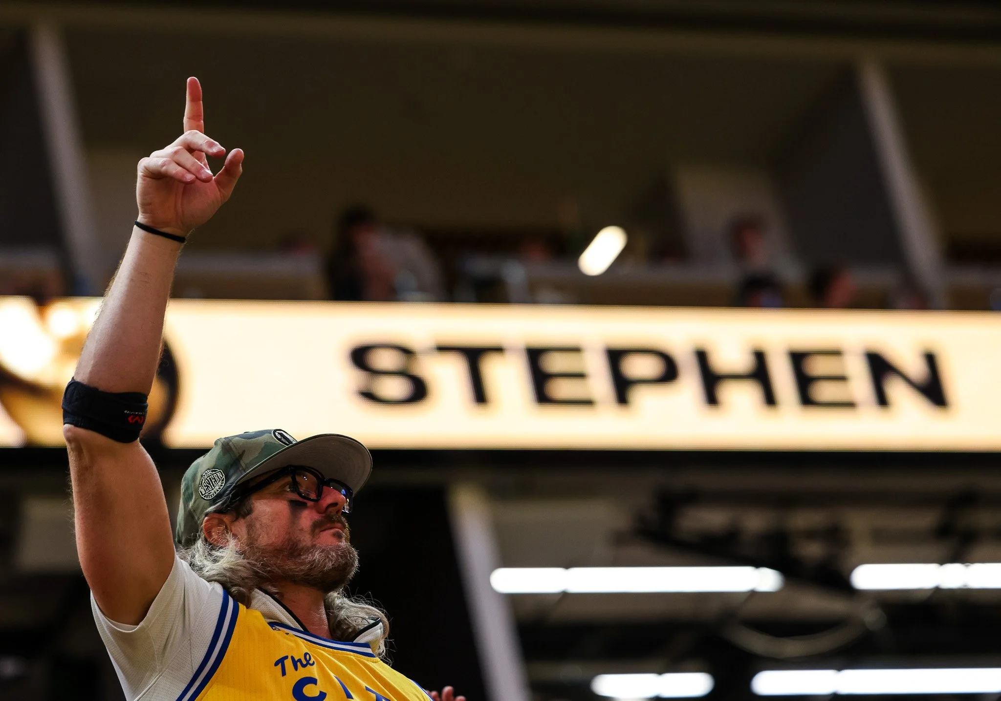 A person with long gray hair, beard, glasses, wearing a baseball cap and a sports jersey, raising their hand with their index finger pointing up. In the background, a sign reads 'STOP' and 'S.'