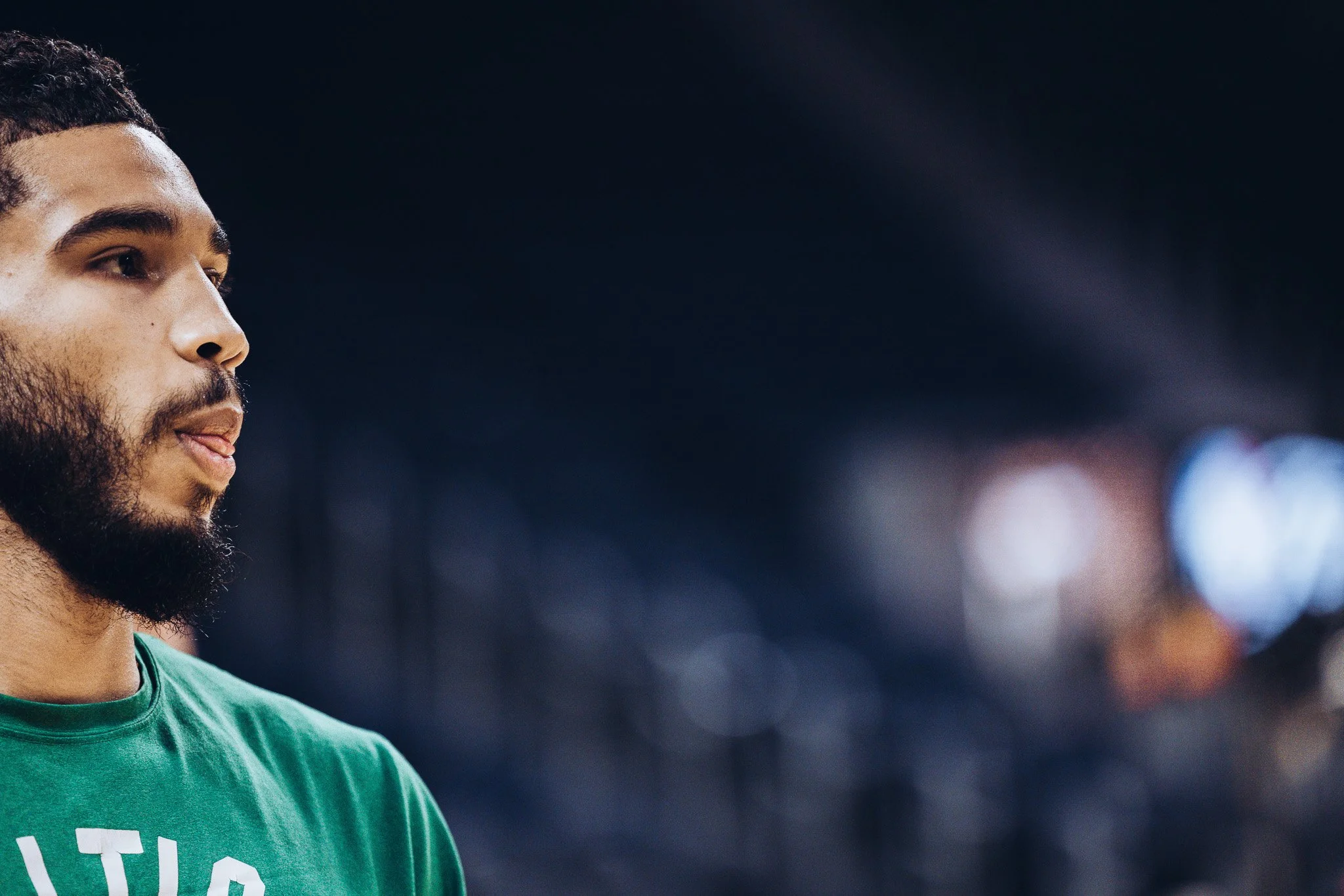 Close-up of a man with a beard and short curly hair, wearing a green shirt, looking focused in a sports stadium with blurred spectators in the background.
