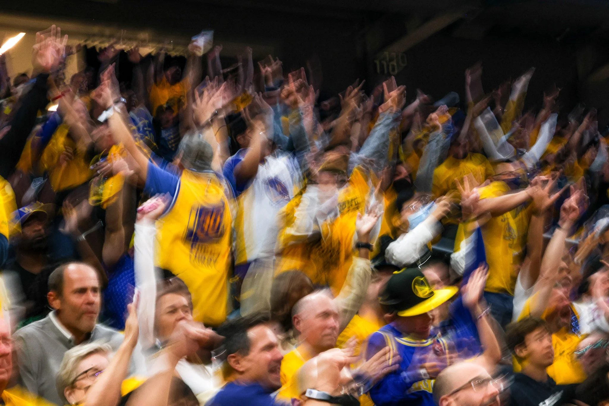 Crowd of people at a sporting event, wearing yellow and blue jerseys, raising their hands in celebration.