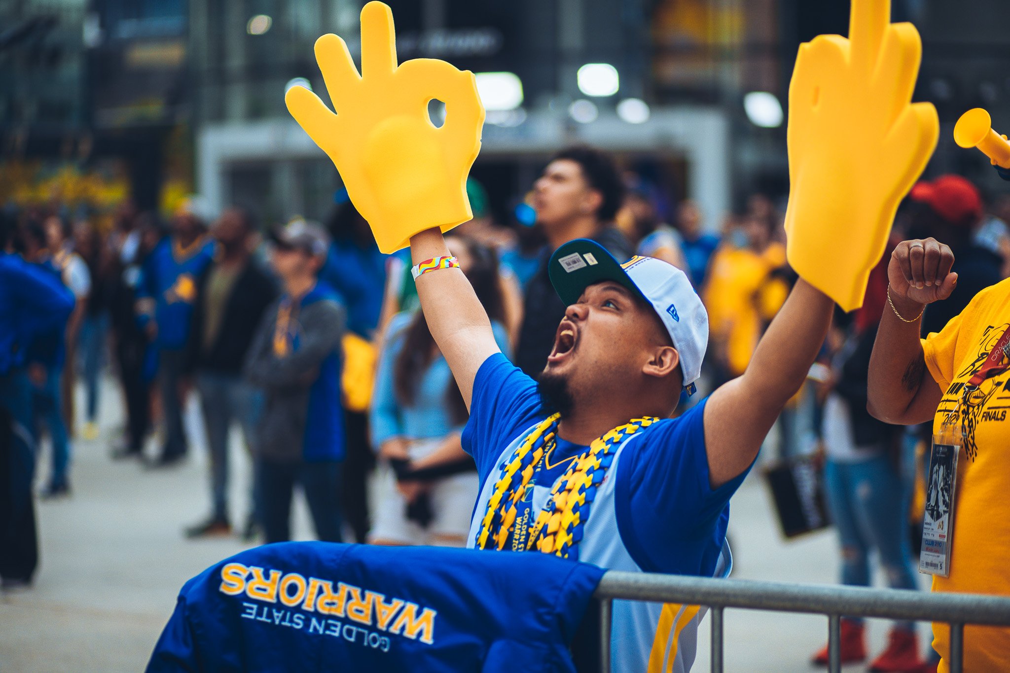 A group of people, including a man in a wheelchair wearing a blue shirt and a white cap, celebrating at an outdoor event. The man is wearing large yellow foam gloves shaped like the "OK" hand sign and raising his arms.