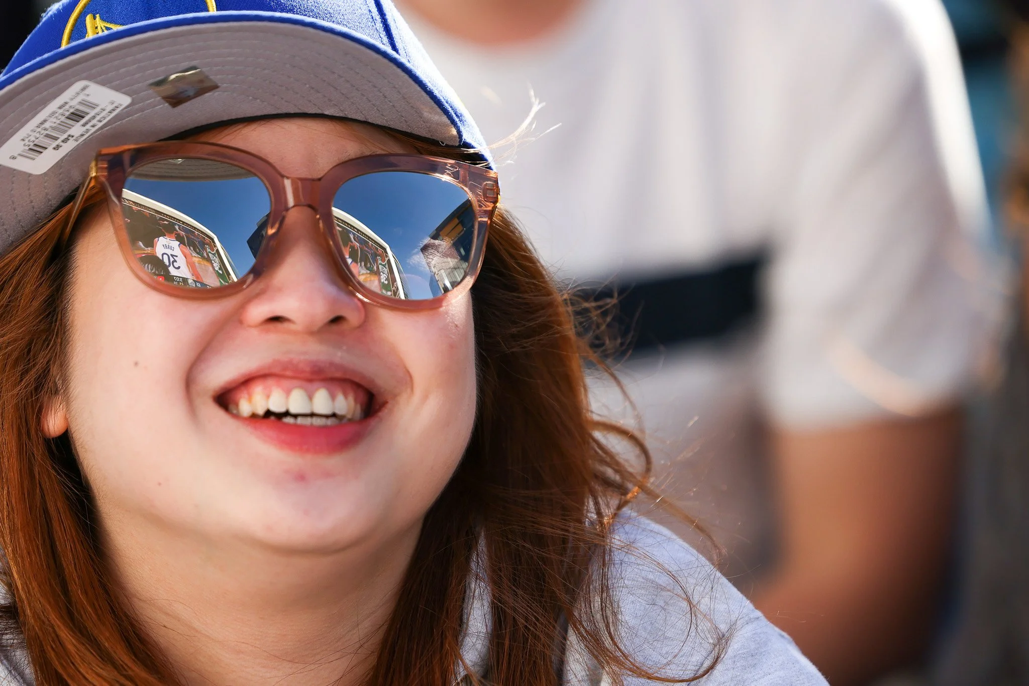 A close-up of a young woman with red hair smiling, wearing pink sunglasses and a gray cap, with reflections of a basketball game in her sunglasses.