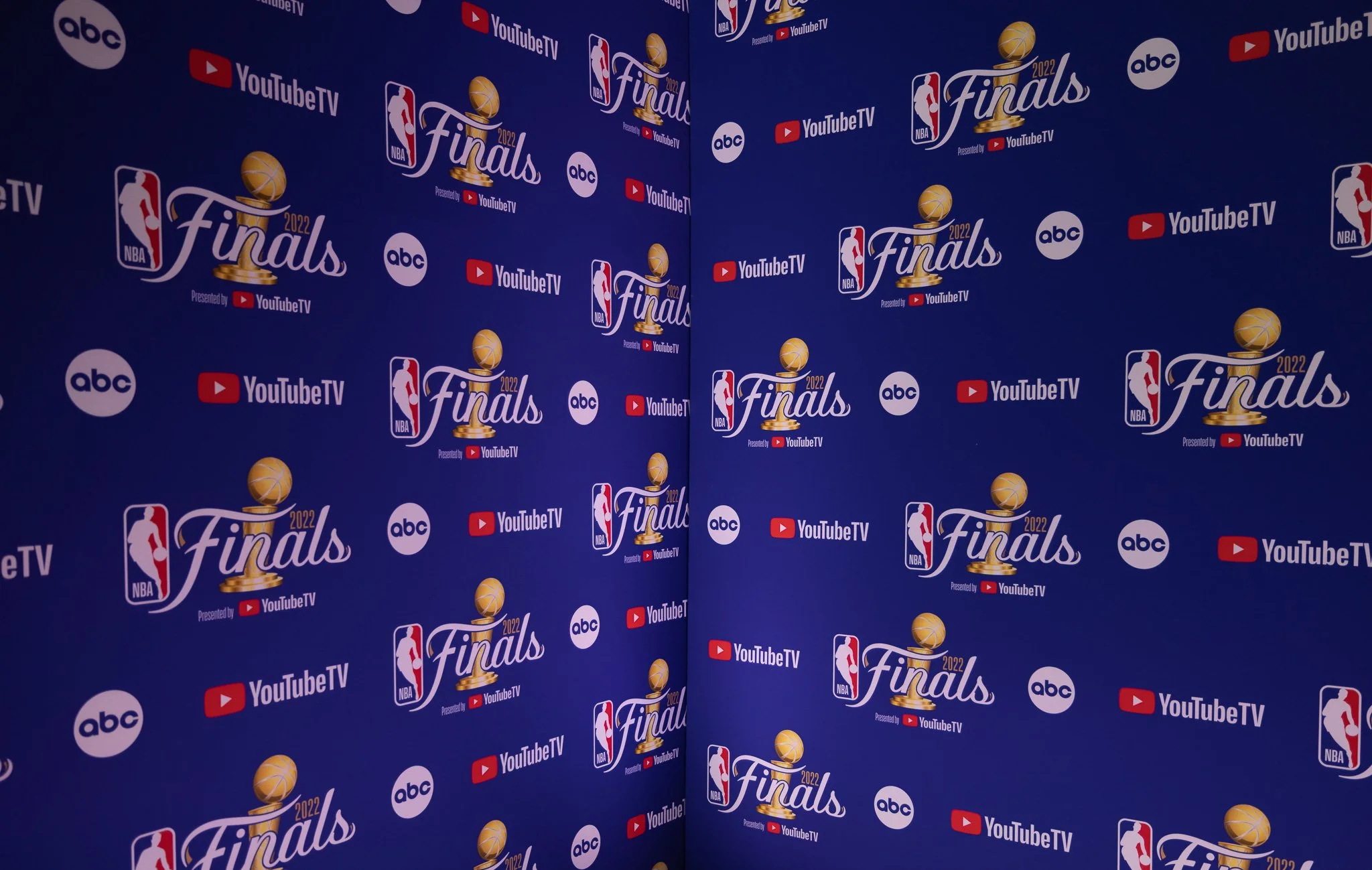 Blue backdrop with logos of NBA Finals 2022, ABC, and YouTube TV, featuring a basketball trophy with the NBA logo and the word 'Finals' repeatedly printed.