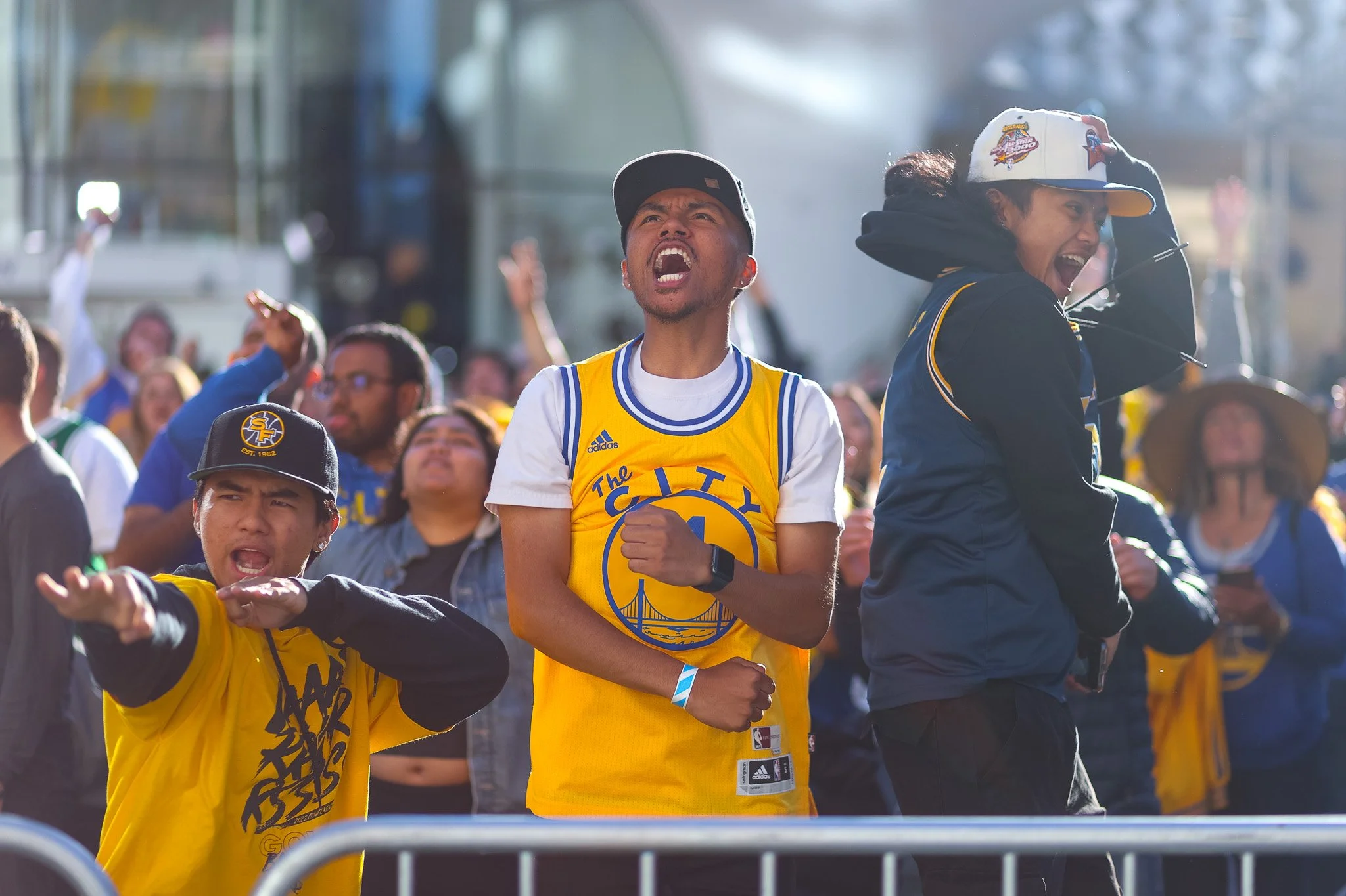 Crowd of people wearing Golden State Warriors apparel, some with their fists raised, celebrating at an outdoor event on a sunny day. Two men are prominently seen in the foreground, one in a yellow basketball jersey and the other in a hoodie and cap, 