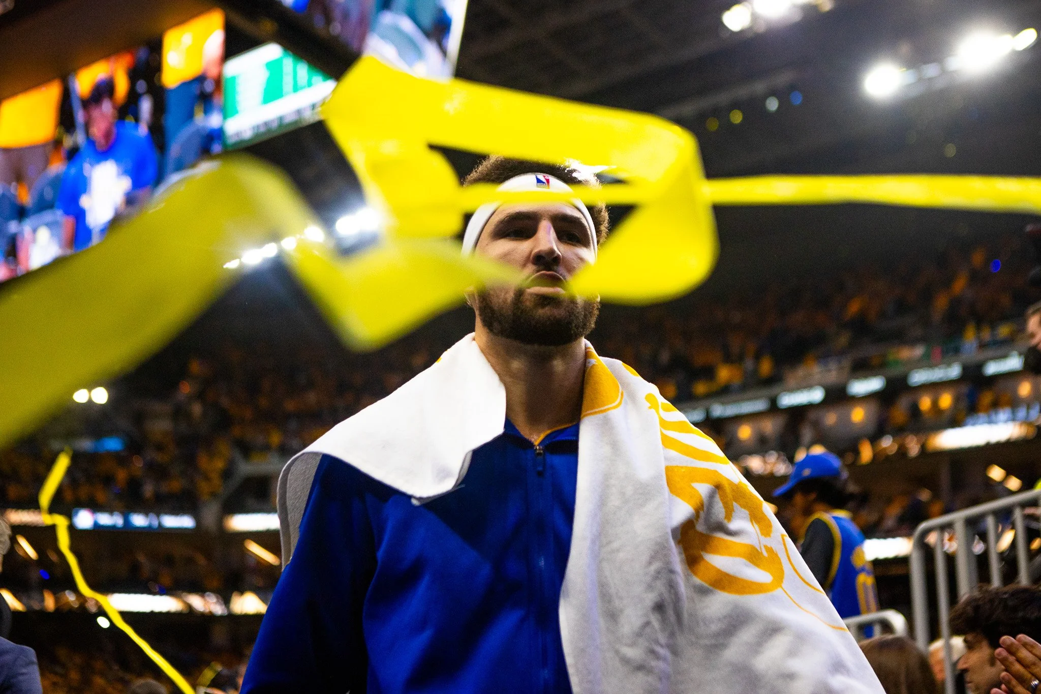 A man with a beard and curly hair wearing a white headband and a blue jacket, seen through a yellow barrier at a sports event, likely football, with a stadium and spectators in the background.