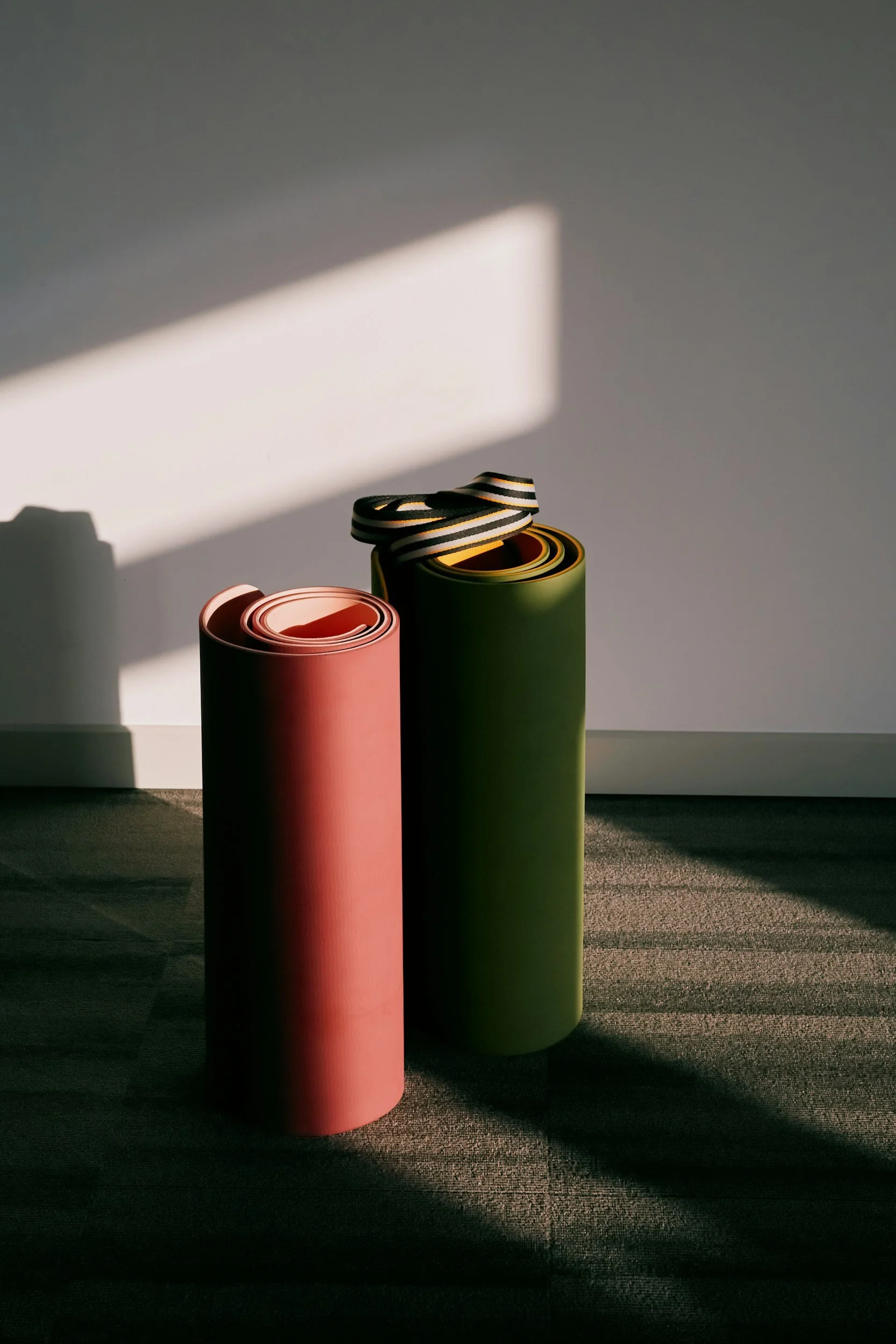 Drei aufrechte Yogamatten in Pink und Grün mit einem gestreiften Yoga-Transportband auf einer glatten Oberfläche, beleuchtet durch Sonnenlicht, das Schatten auf die Wand wirft.